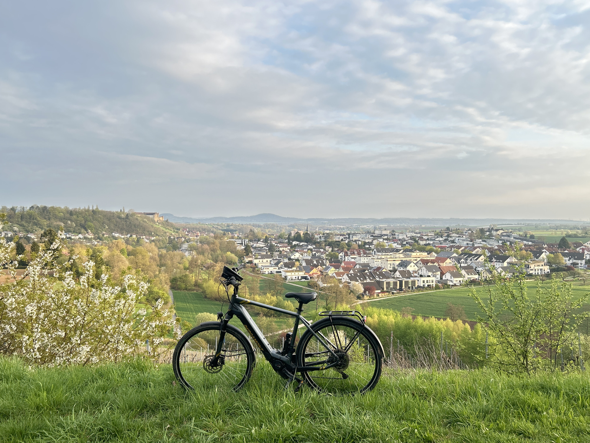 A gray electric bicycle is parked on a grassy hill overlooking a town. The town consists of houses with primarily white walls and red roofs, interspersed with green fields and trees. In the background, a hill with a structure resembling a castle is visible. The sky is cloudy. The location is Unknown, Unknown.