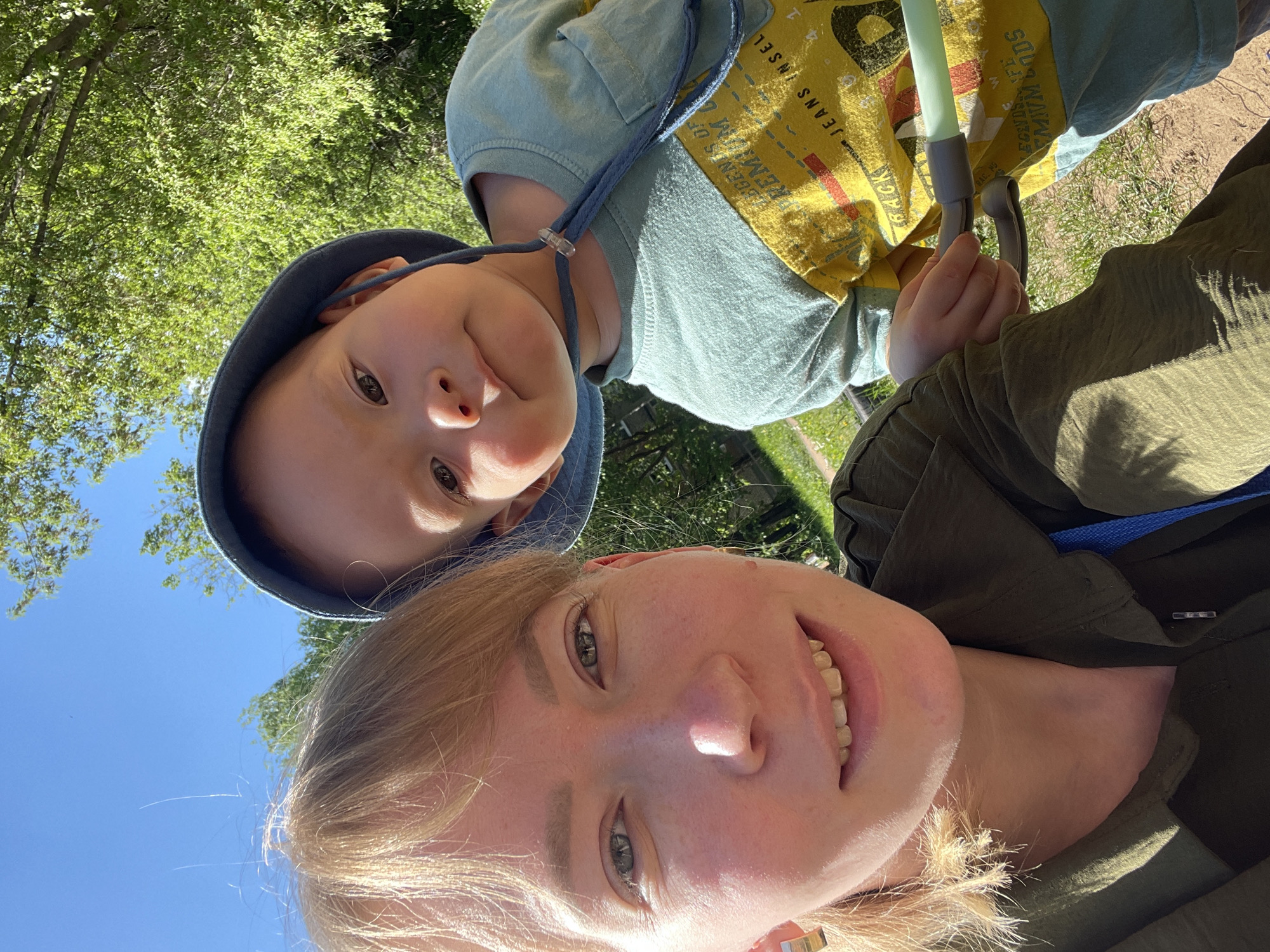 The image depicts a selfie taken by a woman with a young child. The woman has fair skin and blonde hair, and is smiling. The child, positioned above the woman, has fair skin, brown eyes, and wears a blue hooded outfit with a yellow and light blue shirt. They are both holding onto a light green and grey object. The background shows green foliage and a blue sky, suggesting an outdoor location. Without more information, it's impossible to verify if this is in the city of Perm, Russia.