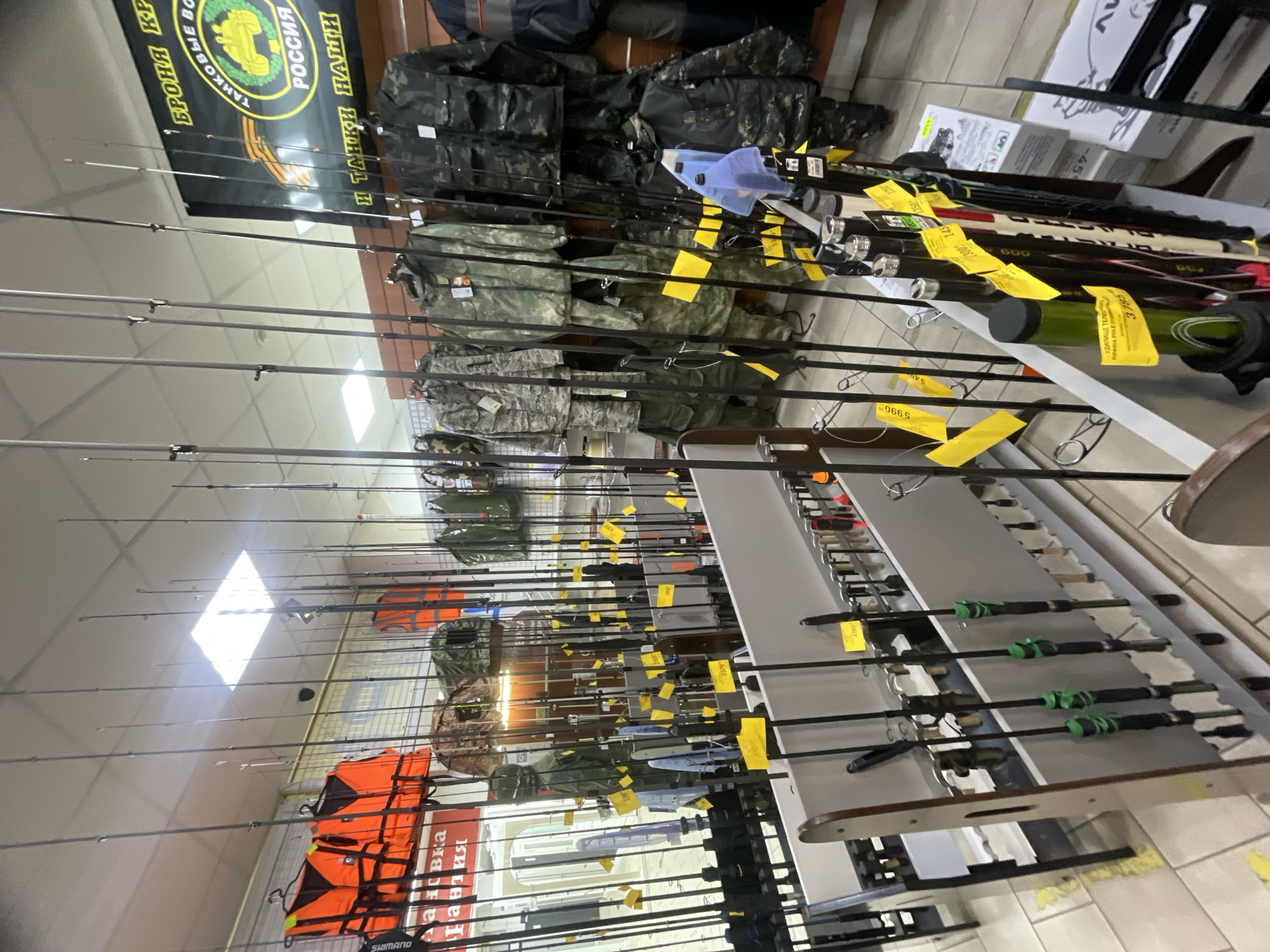 This media file captures an indoor retail setting, likely a store in Krasnoyarsk, Russia, specializing in outdoor gear, fishing equipment, and military-style apparel. The image is taken from a slightly high angle, looking down a narrow aisle lined with products.
The primary subjects are numerous fishing rods, displayed vertically along a tall rack that dominates the left and center of the frame. Many of these rods have small, bright yellow price tags attached. On the right, more fishing rods are arranged horizontally on a multi-tiered display stand. In the background, a variety of camouflage-patterned jackets and pants are visible on hangers, suggesting a selection of hunting, outdoor, or military-style clothing. Bright orange life vests are neatly stacked on a rack towards the bottom left of the image. The store appears well-stocked and brightly lit by overhead fluorescent lights, reflecting off the ceiling tiles. No people are visible, indicating the store might be closed or empty at the time of the photograph.
Notable details include a prominent black banner near the top left corner featuring a tank emblem. The banner has Russian text that reads: "БРОНЯ КР" (Armor KR), "ТАНКОВЫЕ ВО" (Tank-...), "РОССИЯ" (RUSSIA), and "И ТАНКИ НАШИ" (AND OUR TANKS), reinforcing the Russian context of the store. Various yellow price tags are visible on the fishing rods and other items. Some readable prices include "3570", "5990", "1300", "145", "600", "459", and "3785". One tag explicitly identifies an item as "УДИЛИЩЕ ТЕЛЕСКОП" (TELESCOPIC FISHING ROD) and "RIPARIA POLE (CO", likely referring to a brand or material. The brand "SHIMANO" is partially visible near the life vests.