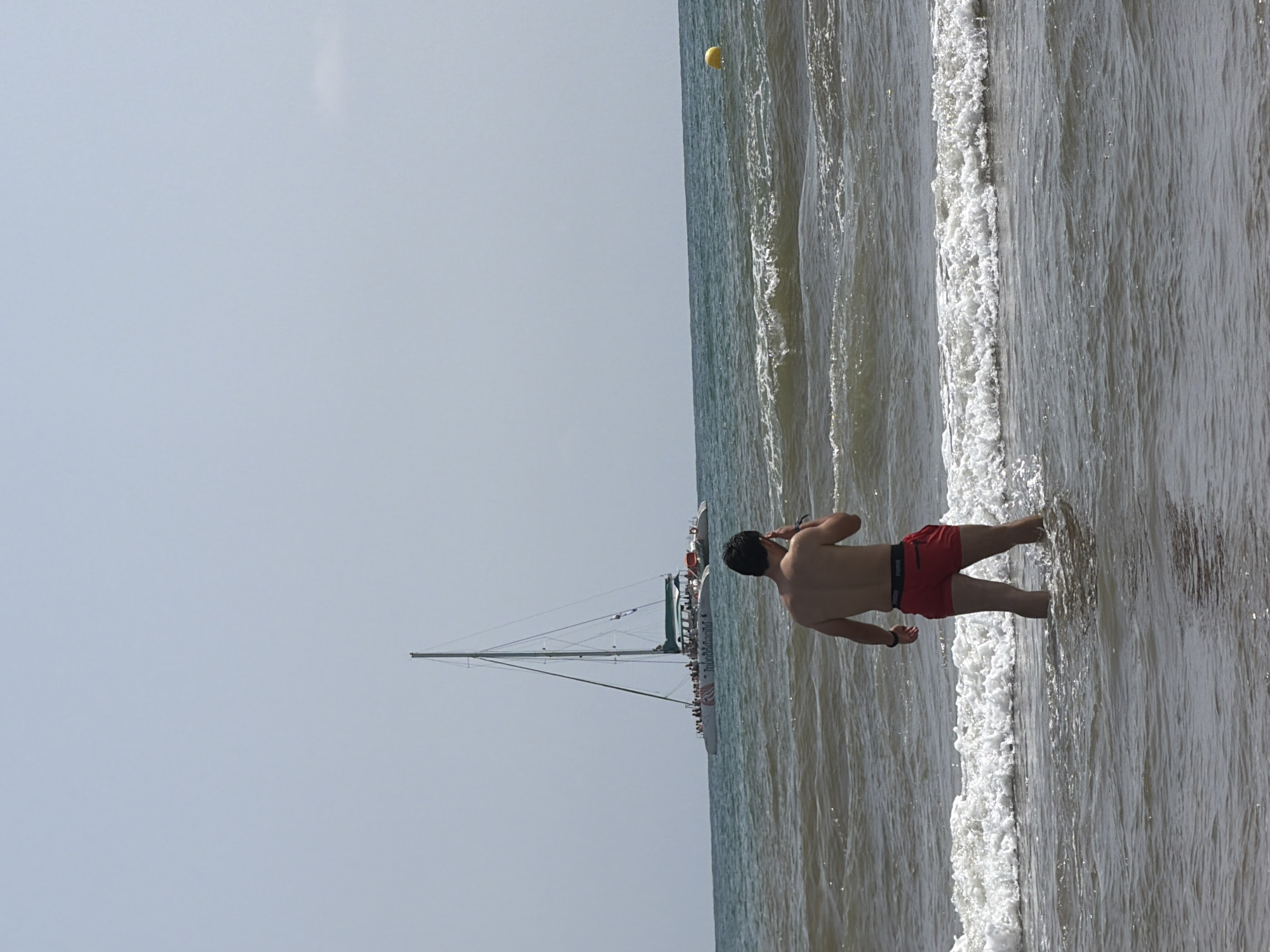 A man is standing in the shallow water at a beach, facing away from the camera. He is wearing red swim trunks. In the background, a catamaran with many people on board is sailing on the ocean. The sky is clear and the water is choppy with small waves. There is a yellow buoy in the distance.