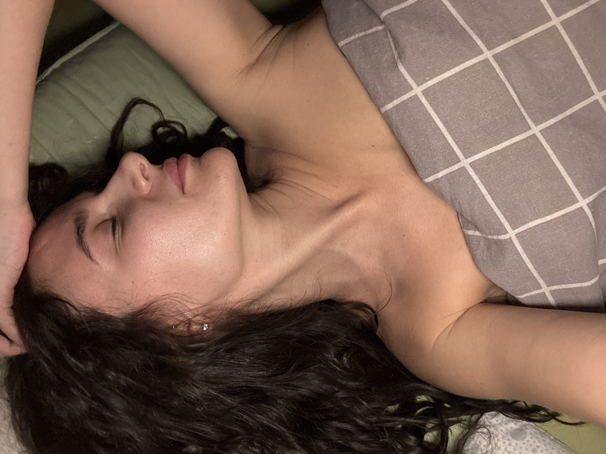A woman is lying down, facing left. She has dark, curly hair and is partially covered by a grey blanket with a white grid pattern. Her right arm is raised. The background appears to be a green pillow or bedding. The location is not discernable from the image.