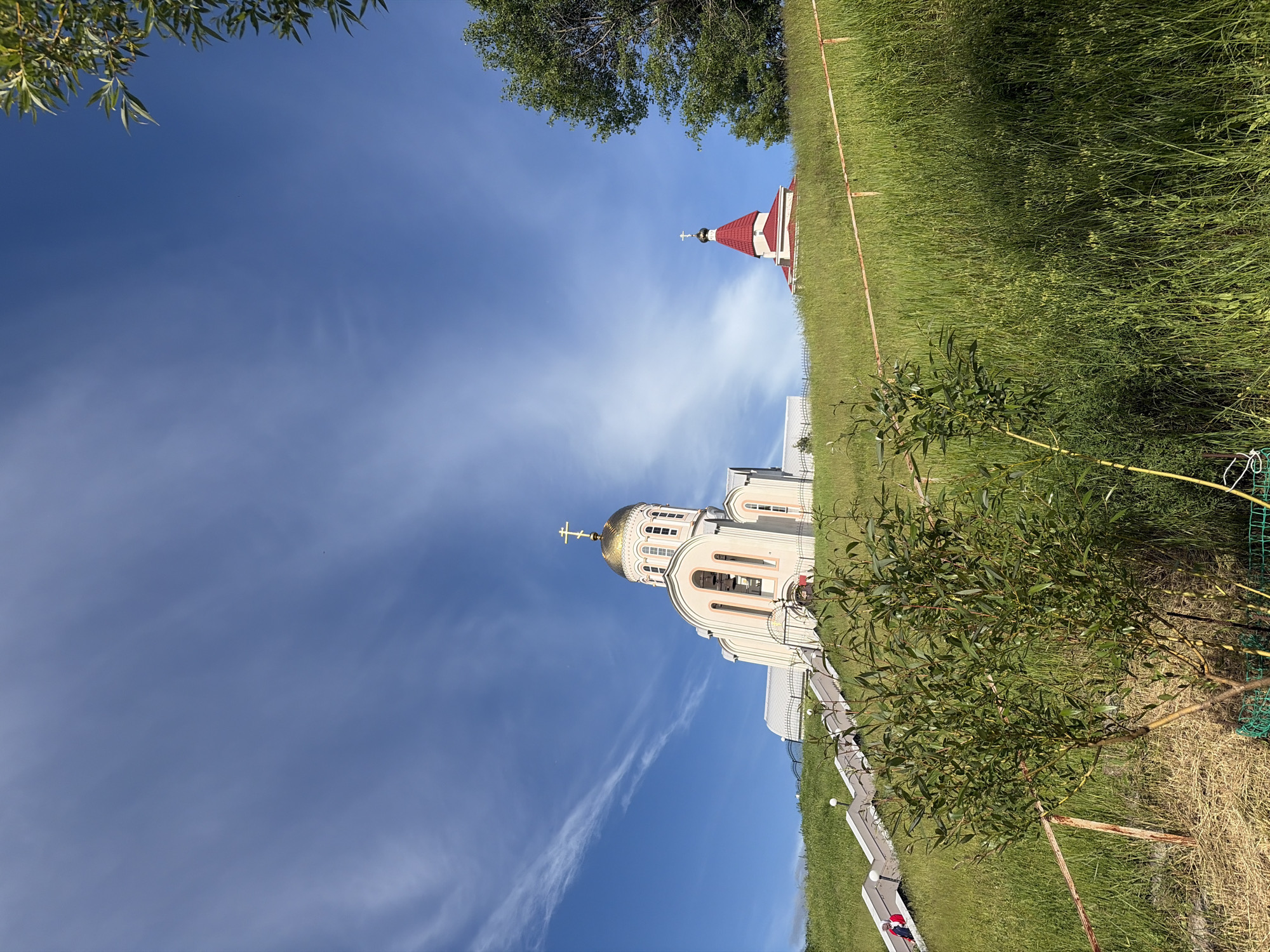 The image presents a serene outdoor vista dominated by two Orthodox Christian churches under a bright blue sky. The main subject is a substantial, light-colored church, likely cream or pale pink, centrally located. It features a striking golden dome topped with an Orthodox cross, and its facade includes multiple arched windows and architectural detailing.
To the right and slightly behind the main church, a smaller religious building is visible. This structure has a distinctive red pyramidal roof, also surmounted by a cross, providing a contrasting element against the sky.
The landscape consists primarily of lush green grass, suggesting either a natural hillside or a well-maintained lawn, encompassing the churches. In the upper left corner of the frame (when oriented correctly), the green foliage of a tree extends into the sky, while a smaller, leafy bush occupies the lower right foreground. A light-colored pathway or a series of steps ascends towards the main church on the right side, flanked by white railings. A green mesh fence is partially visible near the foreground foliage.
The sky is a clear, vibrant blue with scattered, wispy white clouds, indicating a sunny day, likely midday or early afternoon. A small figure in red is faintly visible on the path leading to the main church, but no significant human activity is otherwise depicted. The scene conveys a sense of peacefulness and spiritual tranquility. The architectural style of the churches, with their domes and crosses, is characteristic of Orthodox Christianity, aligning with the specified location of Varvarovka, Russia. No visible text is present in the image.