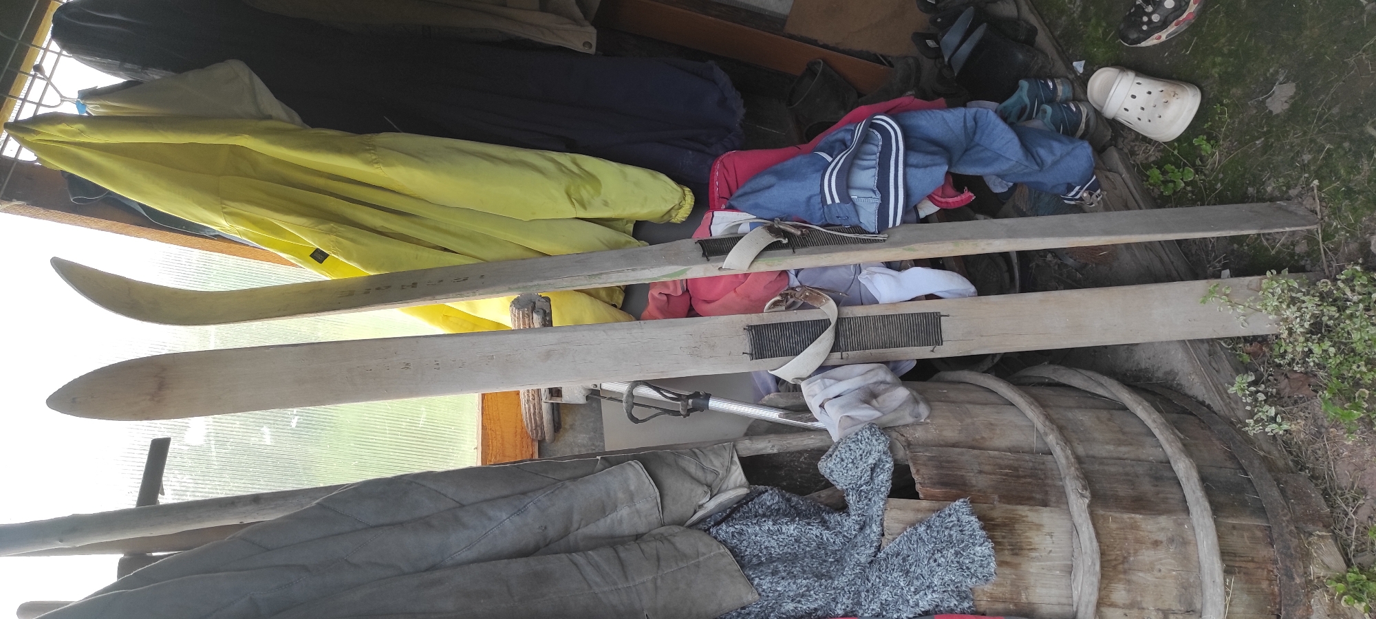 This image shows a collection of items stored in what appears to be an outdoor shed or storage area in Perevoznoye, Russia. The scene is cluttered with various objects, including clothing, skis, and what looks like a wooden barrel.
The main subjects are the objects themselves, piled together. A bright yellow jacket or windbreaker is prominently displayed, draped over a wooden beam. Below it are two wooden skis, positioned vertically. The skis have bindings attached, made of straps and what appear to be metal grids. There are also piles of other clothing, including a blue and white striped jacket, pink and red garments, and gray quilted fabric. A white clog shoe and other footwear are visible in the background, along with dark-colored boots or buckets. The wooden barrel, with its curved staves and metal banding, adds to the rustic feel of the storage area.
The setting is outdoors, as indicated by the natural light and the presence of grass and some small green plants in the lower right corner. The corrugated translucent material visible in the background suggests a greenhouse or a similar structure. The time of day is likely daytime, given the bright lighting, although the specific time is not clear. The weather appears to be mild, as no rain or snow is evident.
There is no discernible activity or event taking place; the image captures a static scene of stored items. There are no people visible in the frame. Notable details include the texture and wear on the wooden skis and barrel, suggesting they have been used or stored for some time.
No visible text is clearly legible in the image.