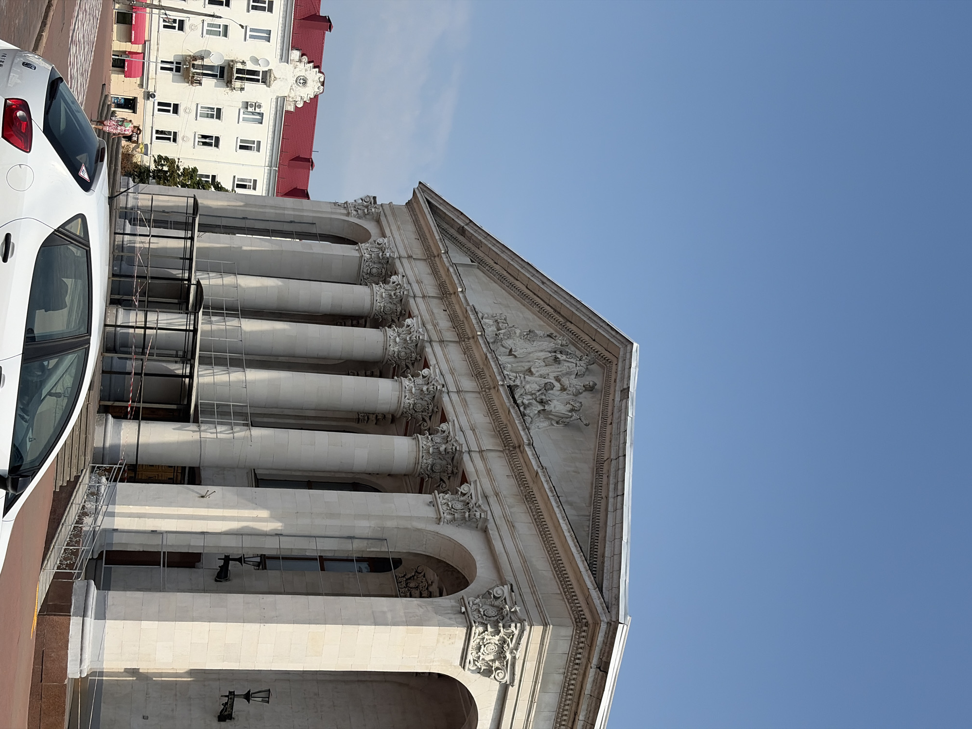 A low-angle, medium shot captures the facade of a grand classical building with a pediment and columns, its light-colored stone contrasting with the clear blue sky. To the left, a white building with a red roof features multiple windows and balconies, and a person in a patterned dress walks on the sidewalk. In the foreground, the rear of a white car is visible. The scene is bathed in daylight, casting shadows on the architecture.