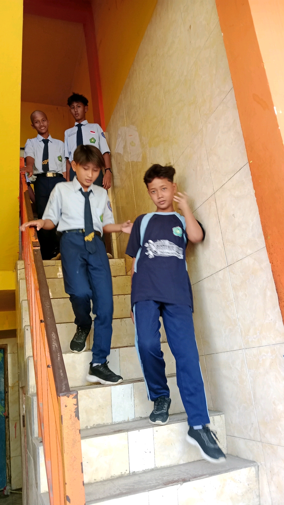 In Waru, Indonesia, four young males, likely students, are descending a light-colored tiled staircase. The staircase features a bright yellow wall on the left and an orange wall on the right, with an orange metal handrail. Two students in the foreground are wearing matching blue pants and dark blue t-shirts with light blue accents and white lettering. The student on the right is looking towards the camera and has his right hand near his face. The student to his left is wearing a white collared shirt, a dark blue tie with yellow accents, and dark blue pants. He is looking down the stairs. Behind them, two other students are visible further up the stairs. One is wearing a similar uniform to the student in the mid-ground, with a white shirt and a dark tie. The other student, closest to the top of the stairs, is wearing a similar uniform and has a shaved head. The lighting suggests it is daytime, possibly indoors or in a shaded outdoor area.