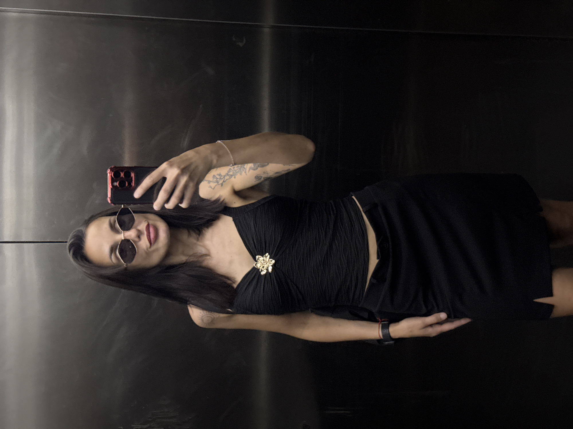 A woman is taking a selfie in an elevator with a reflective metal wall. She is wearing a black dress with a gold flower detail, sunglasses, a watch, and a bracelet. The background is a dark, reflecting the dark outfit and making it difficult to precisely make out the details. The location is unknown and has no indication of being in Pleven, Bulgaria.
