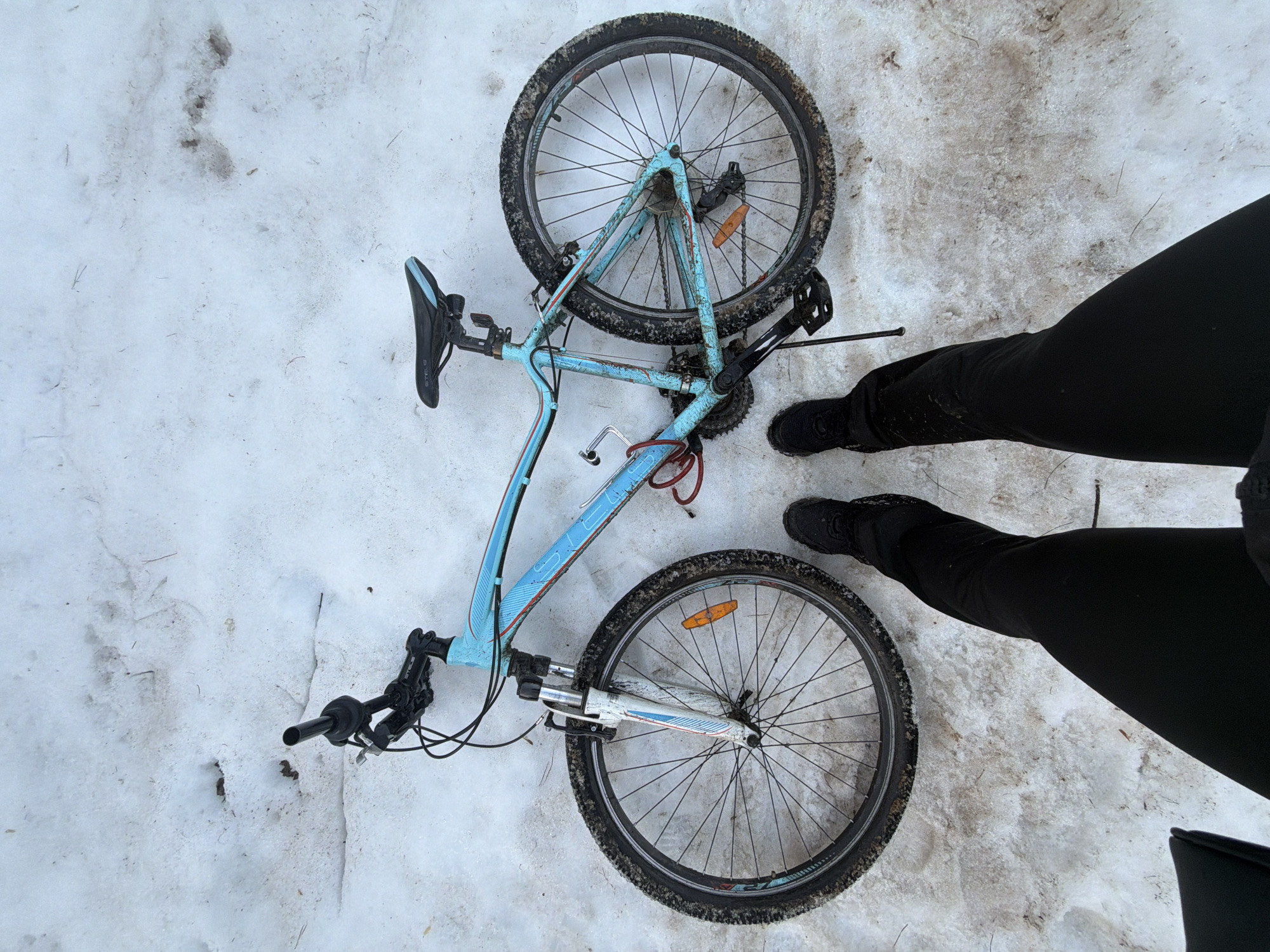 A light blue bicycle lies on its side in the snow. The bicycle's tires appear muddy and the frame shows wear and tear. Next to the bicycle are the legs of a person, wearing black pants and black shoes. The location appears to be snowy outdoor location. There is no evidence in the image to determine its exact location in Chaikovsky, Russia.