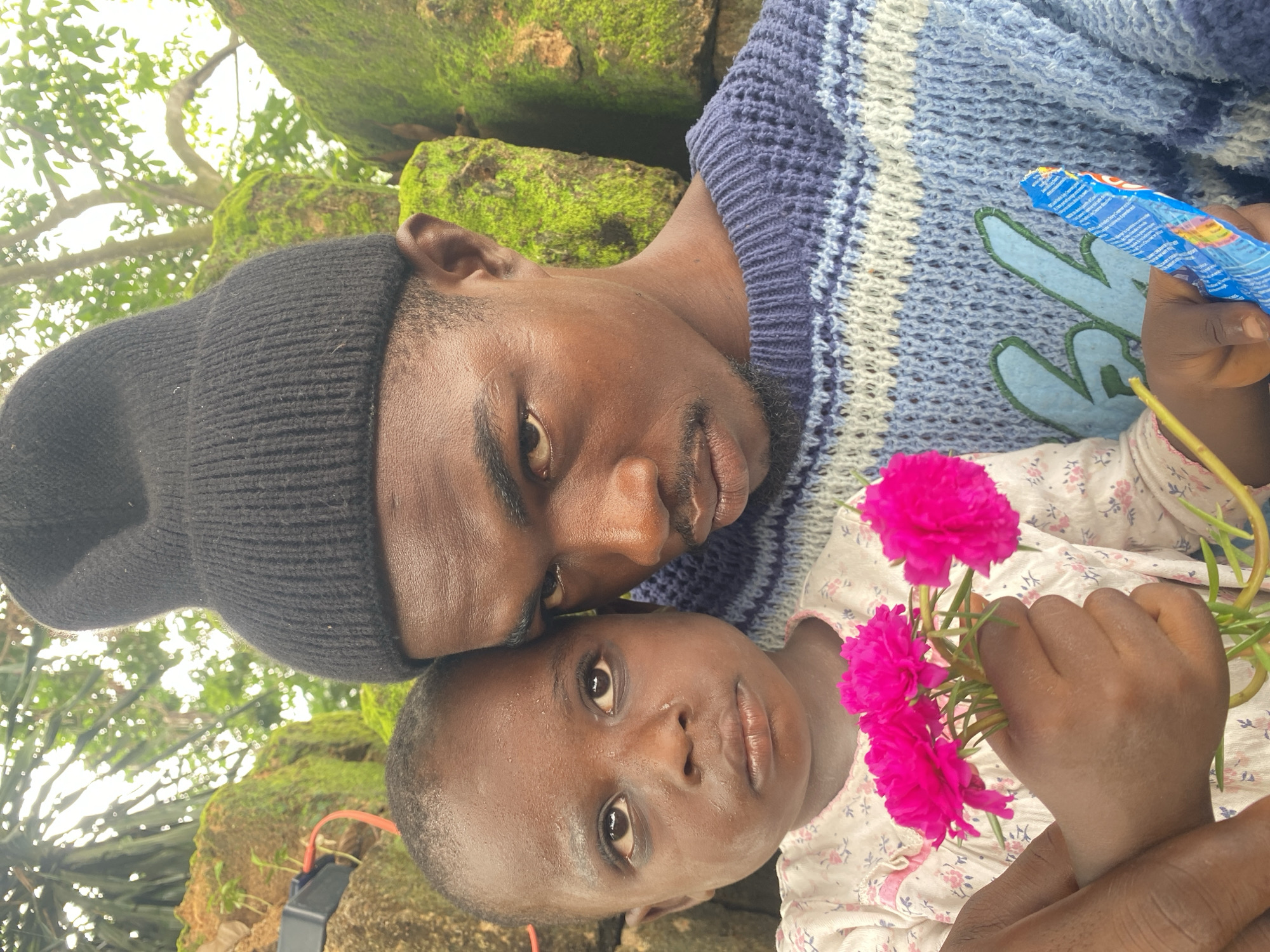 In Igbara, Nigeria, a medium shot captures a man and child lying together outdoors. The man, wearing a black beanie and a blue and white patterned sweater, looks toward the camera. The child, wearing a floral patterned shirt, holds a bouquet of pink flowers. A blue snack wrapper is also visible. Mossy rocks and tree branches with green foliage form the background.