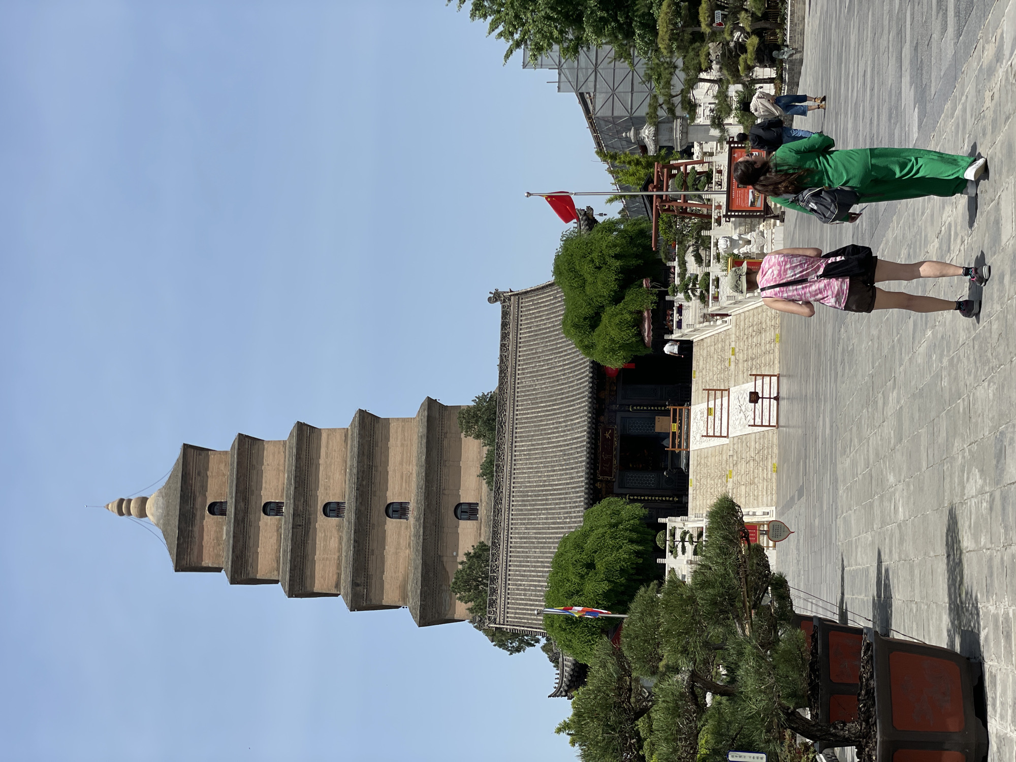 The image shows a view of the Great Wild Goose Pagoda, a Tang Dynasty era pagoda in Xi'an, Yanta District, China. The pagoda is a large, tiered structure with a beige or light brown color. It has several small windows on each level. The sky is a clear, light blue.
In the foreground, there are two people walking on a paved area, possibly a plaza or courtyard. One person is wearing a green outfit, and the other is wearing a pink shirt and shorts. There are trees and other foliage around the base of the pagoda and in the foreground. A Chinese flag is visible. There is also an ornate building next to the pagoda.