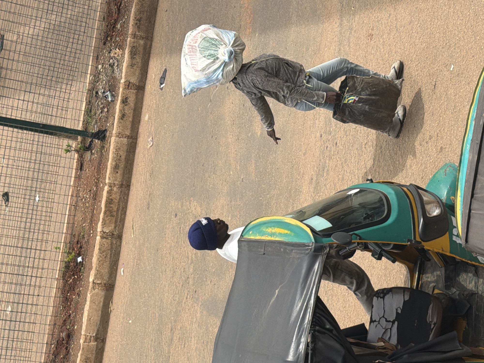 The media file depicts an outdoor scene on a dusty, paved surface, likely a road, in Bukuru, Nigeria. The image is captured from a high angle and is rotated, giving the impression of a tilted perspective.
In the foreground, a green and yellow auto-rickshaw, a common mode of transport in many parts of the world, is partially visible. Its front cab and part of its seating area are in view. A person wearing a blue beanie and a white shirt is positioned within the auto-rickshaw, looking upwards and to the right.
Further back in the image, a person is walking with a large, white bag covering their head. This person is wearing a grey hooded sweatshirt and blue jeans, and their feet are clad in flip-flops. They are carrying a dark-colored bag in their right hand. The presence of a wire fence and a dirt and debris-strewn curb suggest the scene is along a street or pathway, possibly bordering a construction site or an undeveloped area.
The overall impression is of everyday activity in a bustling, possibly hot, environment, indicated by the dusty ground and the casual attire of the individuals. The time of day appears to be daytime, given the clear shadows cast by the auto-rickshaw and the lighting conditions. There are no clear indicators of specific weather conditions, but the dusty environment might suggest a dry period. No visible text is legible in the image.