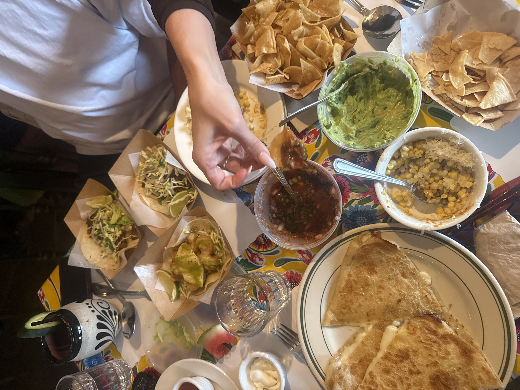 The image shows a table laden with Mexican food, including tacos, quesadillas, guacamole, salsa, and chips. A person's hand is dipping a chip into a bowl of salsa. The dishes are arranged on a colorful tablecloth. The setting may be a restaurant in Los Angeles, United States.