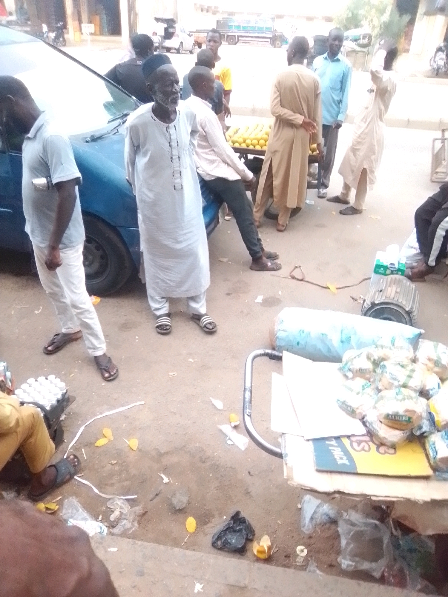 The image depicts a street scene in Arawa, Nigeria, with several individuals and market elements. In the foreground, a person wearing a yellow outfit is partially visible, sitting next to a crate of white items. To the left, a man in a light blue shirt and white pants stands beside a blue car. Further back, an elderly man with a white beard and a white traditional garment is looking to his right. Another man in a similar white garment is standing to his left. A man in a tan robe is facing away from the camera, and another man in a light blue shirt is standing behind him. Several people are milling about in the background, near a table laden with oranges. The ground is dirt and debris, with various items scattered around, including plastic bags, discarded food, and a large metal object that appears to be an industrial motor. A cart with items is visible on the right side of the frame. The overall impression is of a busy outdoor market or gathering.