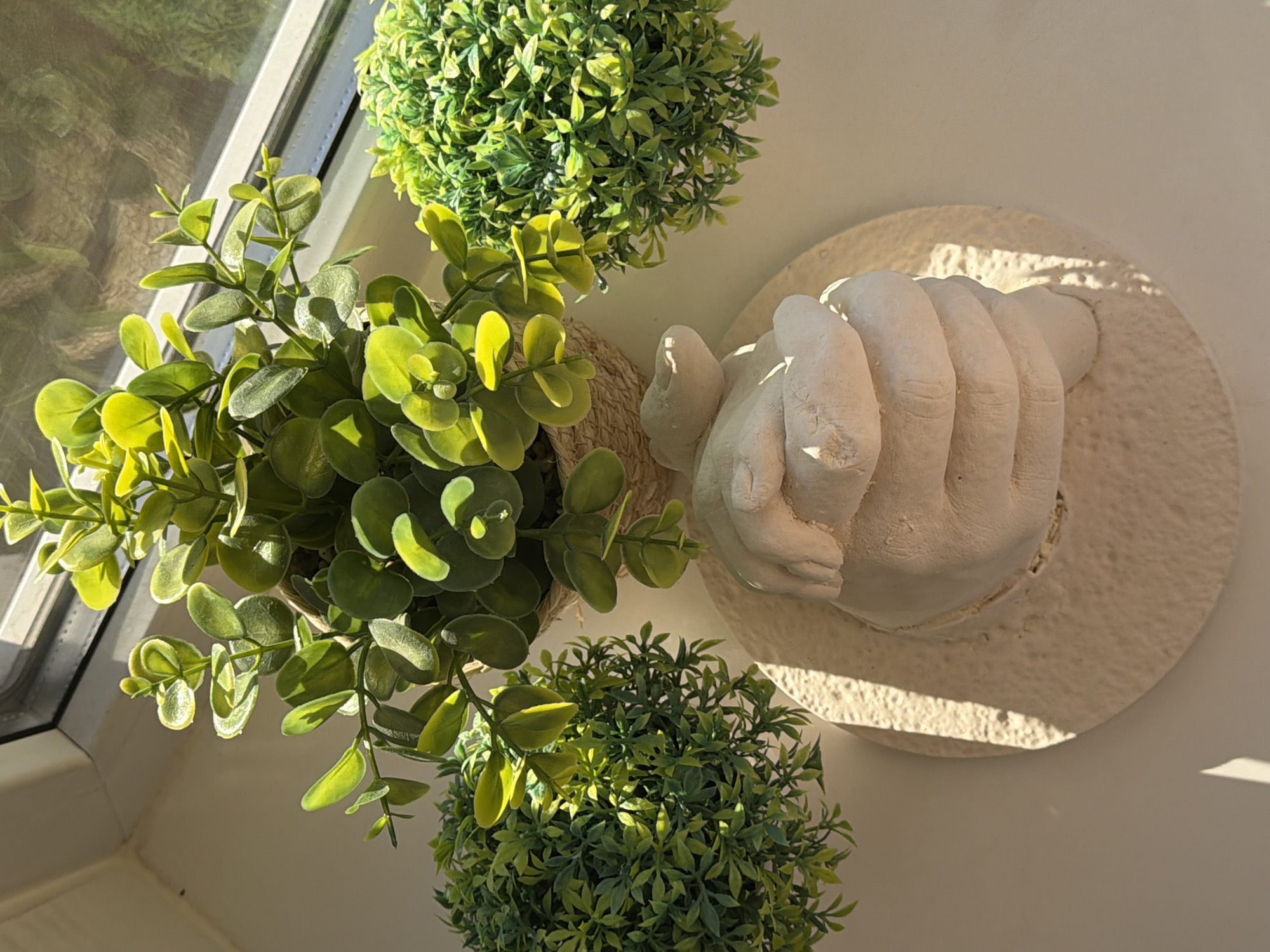 The image depicts a windowsill in a room, illuminated by sunlight. Two artificial green plants in pots sit on the left and bottom, with a third plant positioned in the upper-center. To the right of the center plant is a plaster cast of a hand, mounted on a round, textured plaster base. The windowsill is part of a window with a white frame. The light suggests a bright, sunny day. There is no evidence to confirm the city location.