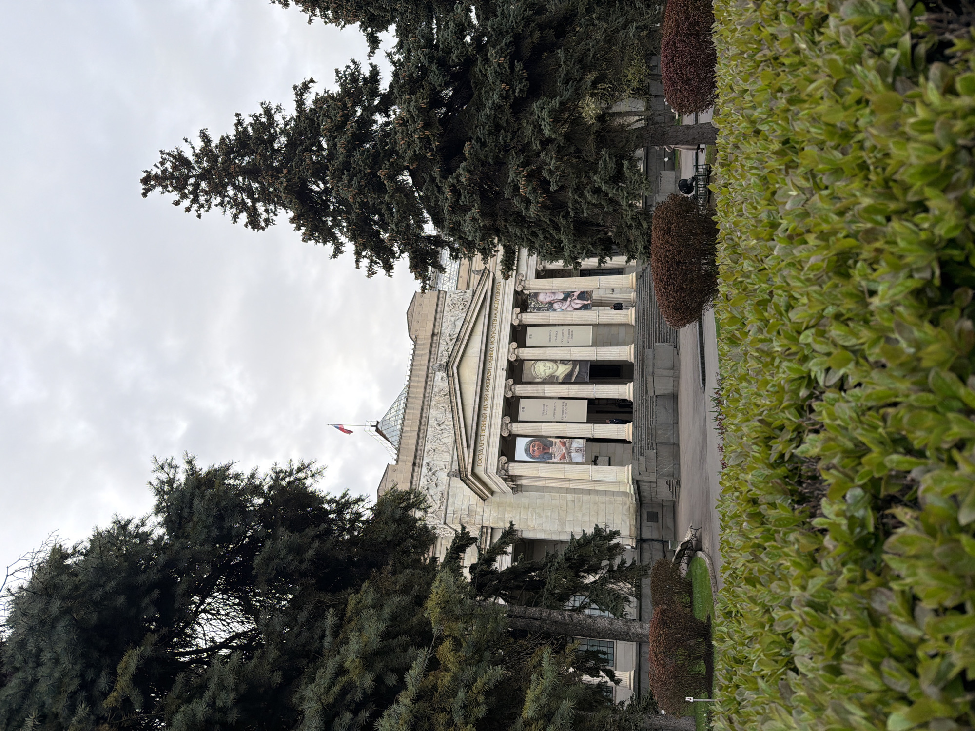 The image captures an exterior view of the State Museum of Fine Arts (Pushkin State Museum of Fine Arts) in Moscow, Russia, seen from a slightly elevated angle. The grand, classical-style building dominates the mid-ground, featuring a stone facade with large columns supporting a pediment adorned with sculptures. A glass dome or skylight is visible on the roof, with a flag (possibly Russian or French, given the colors) flying from a pole.
Banners advertising exhibitions hang between the columns. Visible text in Cyrillic on the building's frieze clearly reads "ГОСУДАРСТВЕННЫЙ МУЗЕЙ ИЗОБРАЗИТЕЛЬНЫХ ИСКУССТВ" (State Museum of Fine Arts). The banners display portraits, including one of an older man in profile and another of a woman, possibly of ancient Egyptian style. Partial Cyrillic text like "ВЫСТАВКА" (Exhibition) and "ГРАФИКА" (Graphics) is discernible on these banners.
The building is flanked by numerous large evergreen trees, some obscuring parts of the facade. In the foreground, a long, well-maintained hedge of vibrant green bushes runs parallel to the building's entrance, with several rounded, reddish-brown trimmed bushes positioned behind it. Two figures, possibly visitors or staff, are visible near the building's entrance, suggesting general public activity.
The scene depicts a cloudy, overcast day, with no direct sunlight. The overall impression is that of a significant cultural institution in an urban setting.