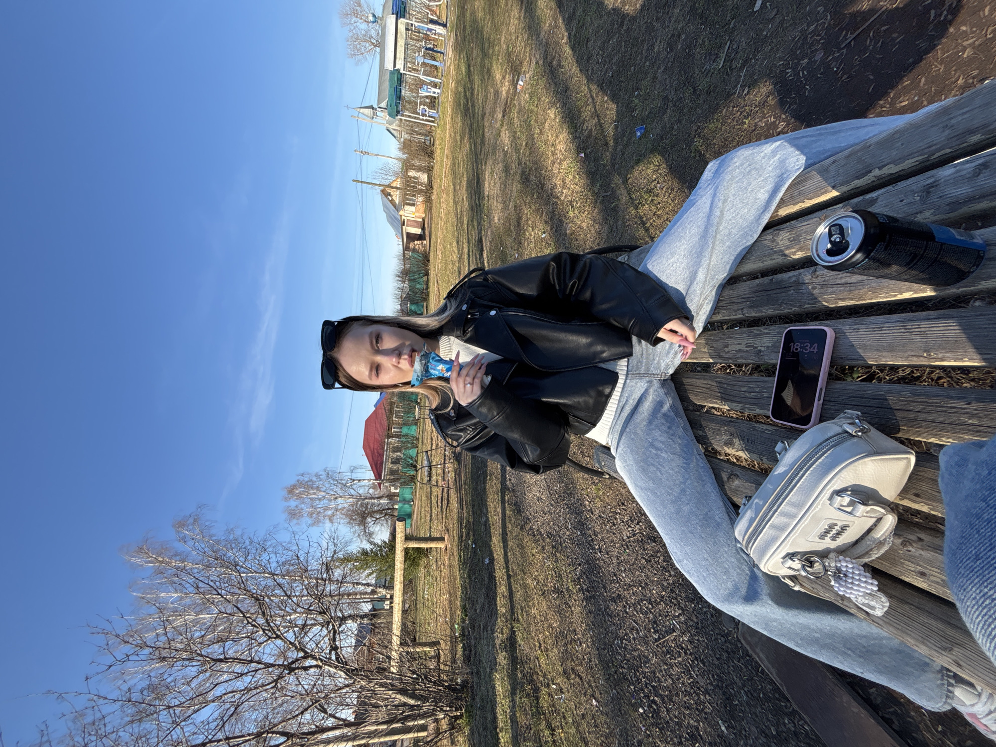 An outdoor image captures a young woman relaxing on a wooden park bench under a clear blue sky, likely in Фоки, Russia. She has light-colored hair, is wearing a black leather jacket over a white sweater, and light blue jeans, with sunglasses pushed onto her head. She holds a blue patterned bottle with a white cap to her mouth, appearing to drink. Her legs are extended on the bench, and her left hand displays light pink fingernails.
Beside her on the bench are several items: an open black energy drink can, a pink smartphone displaying "18:34", and a small light grey handbag with a visible "MIU MIU" logo and a white beaded keychain.
The background shows an open area with dry grass and patches of dirt. Bare trees are present, suggesting early spring before leaves have fully emerged, or late autumn. Distant buildings, including some with red roofs and a small church-like structure, are visible, indicating a rural or semi-urban setting. Strong shadows fall across the ground and bench, indicative of bright sunshine, consistent with the late afternoon time displayed on the phone. The woman's expression is neutral, conveying a calm and relaxed demeanor.