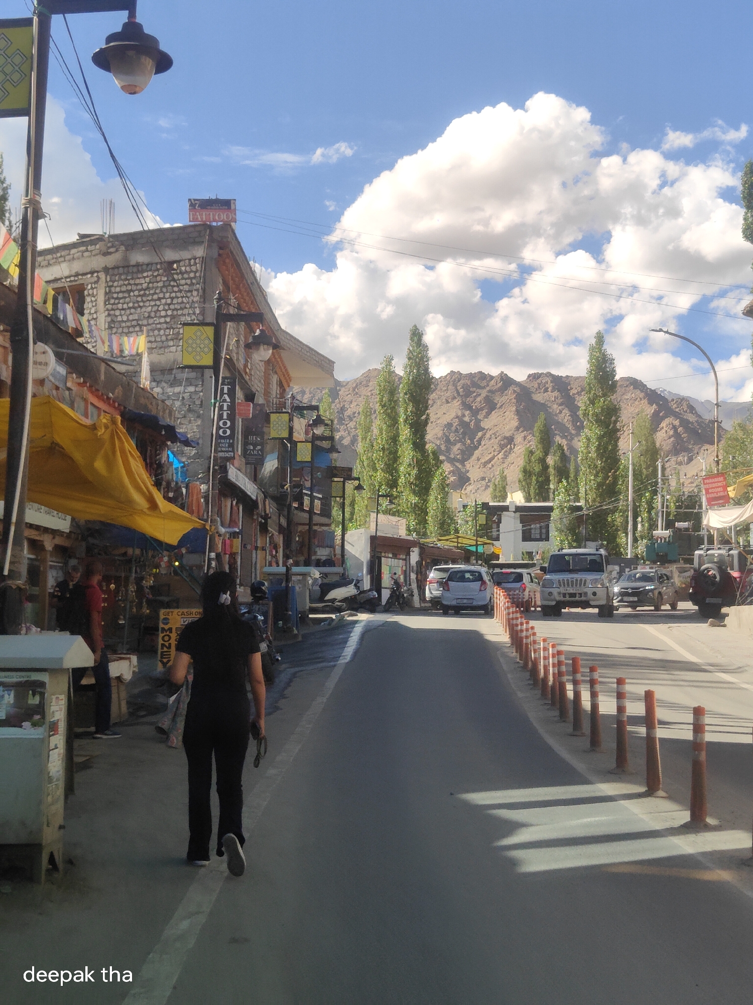 The image depicts a street scene in Leh, India. On the left side of the street, there are shops with various signs, including one for "TATTOO". A woman wearing a black top and black pants is walking away from the camera on the sidewalk. Several cars and a motorcycle are visible on the road. In the background, there are tall trees and mountains under a blue sky with white clouds. The street is lined with orange and white traffic cones. A street lamp is visible at the top left of the image. The overall impression is of a sunny day in a mountainous town.
