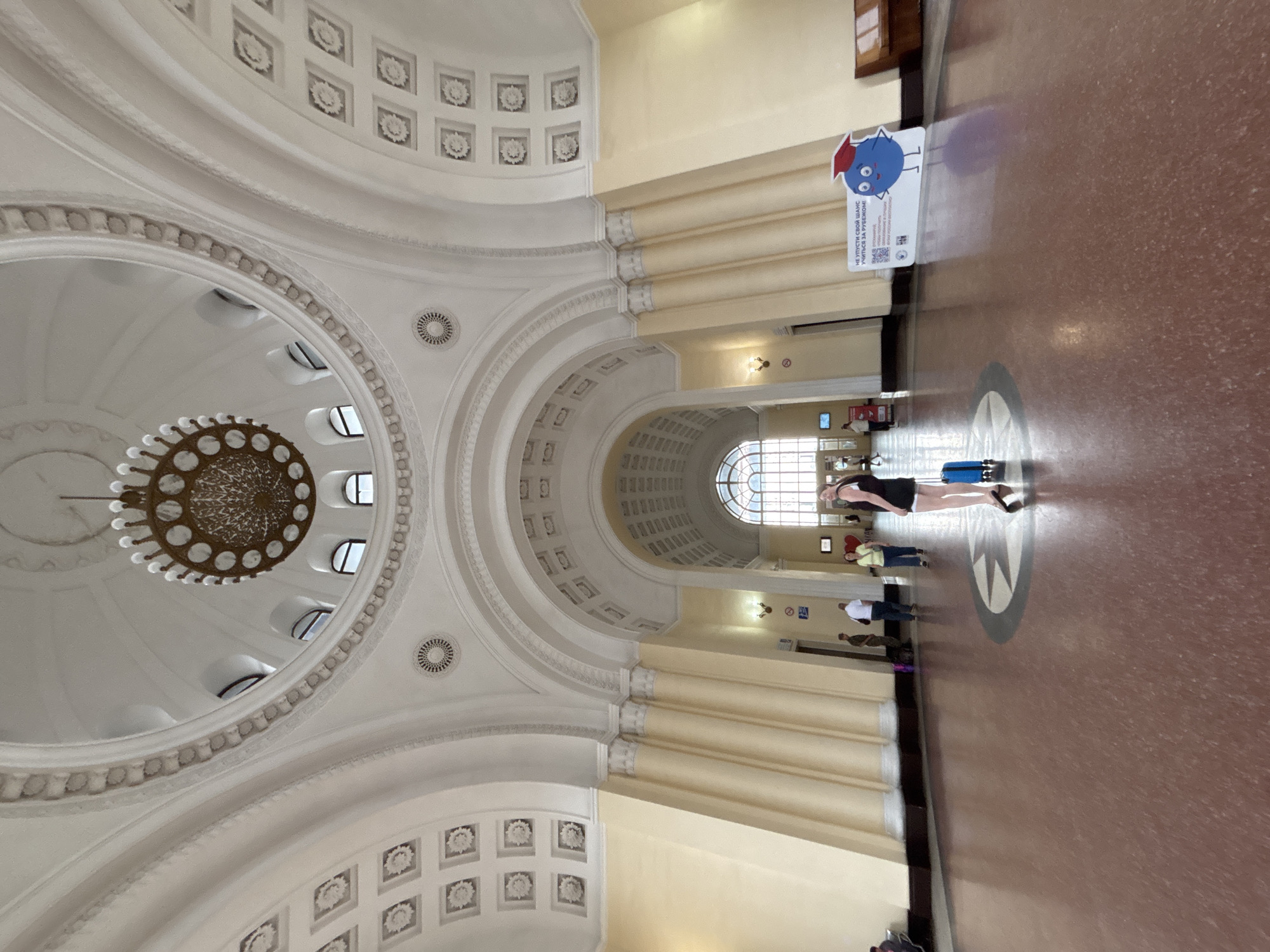 The image depicts the interior of a grand building, likely a train station, with a high, domed ceiling adorned with ornate moldings and a large chandelier. The architecture features large arched doorways and windows, with light flooding in from a window at the far end of the hall. The walls are painted a pale yellow, and the floor is a polished reddish-brown.
In the foreground, a young woman stands with a blue suitcase, facing away from the camera. She is wearing a black top and shorts. Several other individuals are visible in the mid-ground, some walking and others standing in conversation. One individual appears to be in a military uniform. A sign with cartoon graphics and text in Russian is positioned on the right side of the hall.
Based on the architectural style and the presence of the Russian text on the sign, this location is likely the Yerevan Railway Station in Yerevan, Armenia. The station was built in the Soviet era and retains much of its original design.