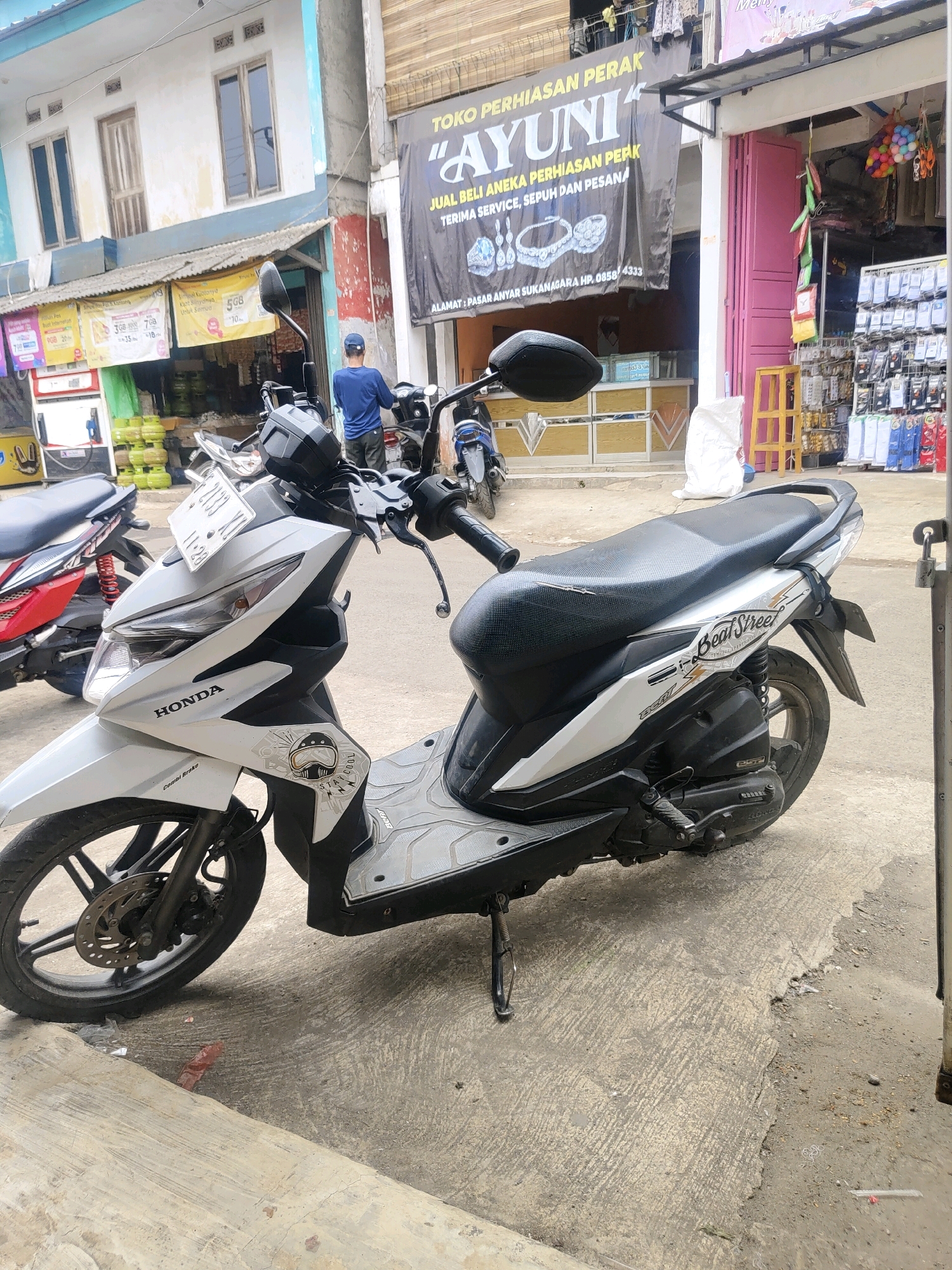 A white Honda Beat Street scooter is parked on a concrete sidewalk in front of a shop in Sukanagara, Indonesia. The scooter is angled to the right, showing its left side. A license plate is visible on the front of the scooter. The shop behind the scooter has a sign that reads "TOKO PERHIASAN AYUNI" and displays images of jewelry. The shop also has a pink storefront and shelves filled with various items, including what appear to be socks. A person wearing a blue shirt and a blue cap is standing in front of the shop, facing away from the camera. Another red scooter is partially visible to the left of the white scooter. The environment appears to be a commercial street with buildings and other parked vehicles.