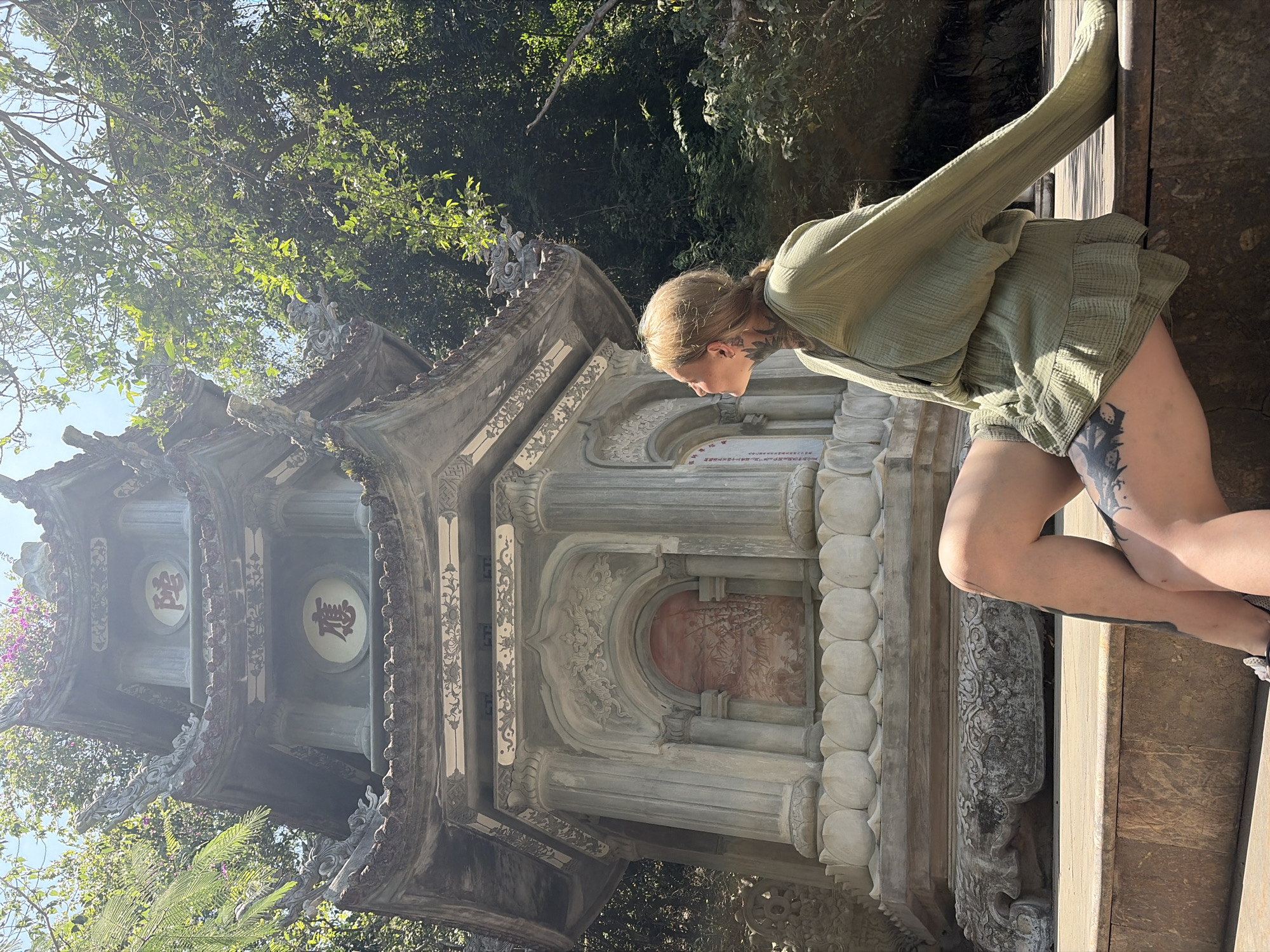 The image captures a young woman next to an ornate traditional structure, likely a pagoda or temple, under bright daylight. The scene is set outdoors with lush green trees and a clear sky visible in the background, suggesting a sunny day. The overall aesthetic is one of cultural exploration and appreciation.
The woman, with light brown hair pulled back, is seen from the side, looking towards the structure. She is wearing a light green, loose-fitting outfit consisting of a long-sleeved top and ruffled shorts. Visible tattoos adorn her neck and upper thigh, adding a personal detail to her appearance. Her posture suggests she is either observing the structure closely or pausing for a photo.
The traditional structure is elaborately decorated, featuring multiple tiers with curved eaves. It is constructed from stone or concrete, weathered to a greyish hue. Notable architectural details include carved dragon-like figures on the roof corners, intricate floral or geometric patterns along its facades, and columns with distinctive round, layered forms. Inside circular panels on the upper tiers, two red Chinese characters are prominently displayed. Another section of the structure features a reddish-brown arched recess with subtle painted imagery, possibly depicting bamboo. The structure appears to be set within a natural, perhaps park-like, environment, consistent with locations in Đà Nẵng, Vietnam. The scene combines historical architecture with a contemporary figure in a serene, sunlit setting.