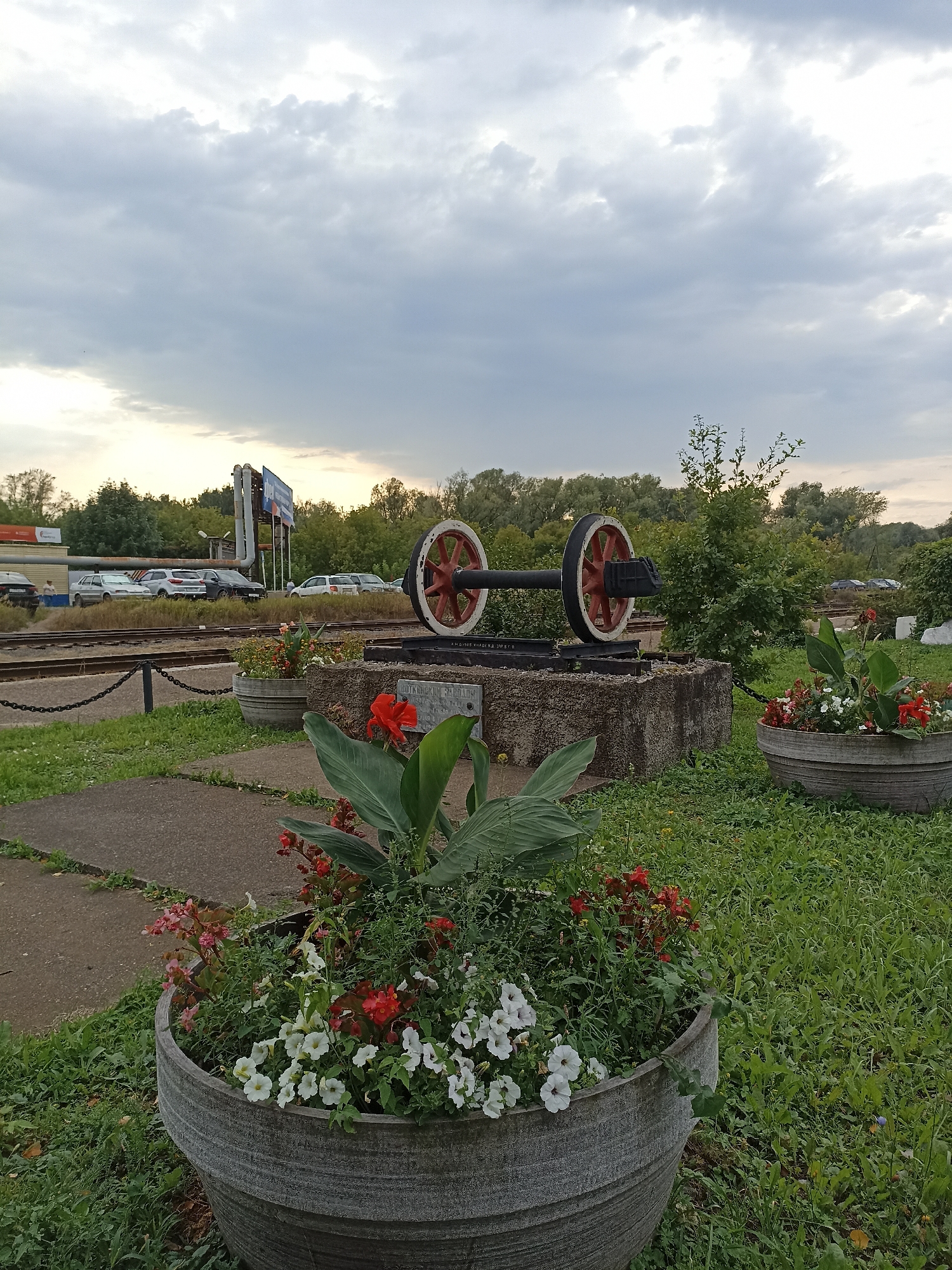 In the city of Votkinsk, Russia, a large, weathered stone planter overflowing with red and white flowers sits in the foreground of the image. Behind it, a monument featuring a pair of large train wheels on an axle is displayed on a concrete pedestal. The wheels have red rims and white hubs. A plaque is visible on the pedestal. Beyond the monument, train tracks run horizontally across the scene, with a low chain-link fence bordering them. Several cars are parked in the background, near some buildings and trees. The sky above is overcast with dramatic, gray clouds.