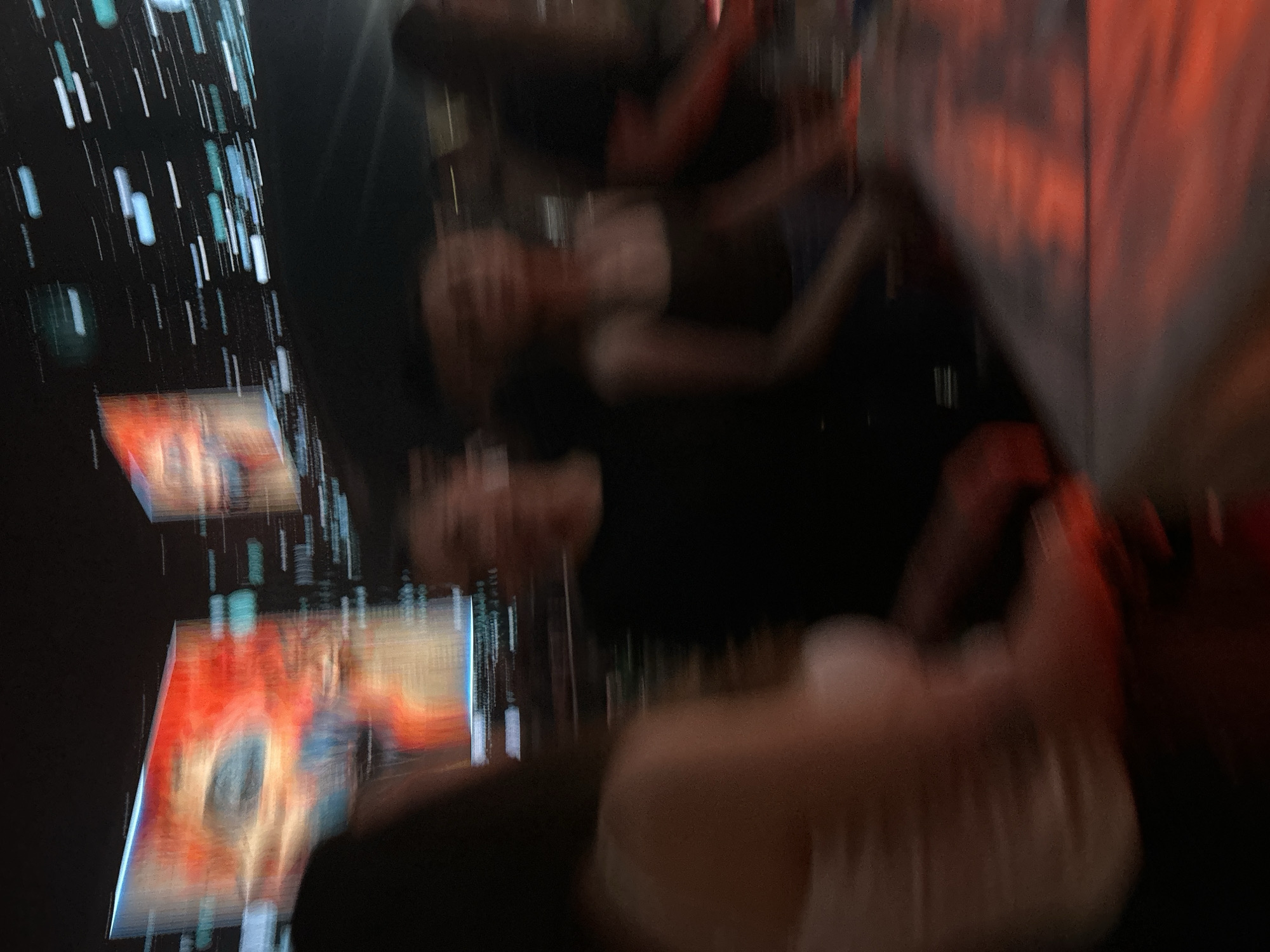 The image captures a blurry, dynamic scene at night, possibly in a lively urban setting. In the background, two large screens display vibrant, abstract images with prominent red and orange hues, suggesting an entertainment venue or a public event. Streaks of light, possibly from moving vehicles or the screens themselves, create a sense of speed and energy.
In the foreground, several individuals are visible, their features blurred by motion. The closest subject is a person in the left foreground, seen from the back, wearing a light-colored top. To their right, another person is visible in a dark top. Further back, two more individuals are partially discernible, their forms indistinct. The overall impression is of a group of people engaged in an activity or observing something in a dimly lit, atmospheric environment. The red light reflecting on the surface in the lower right corner could indicate a bar, restaurant, or stage lighting. Without specific landmarks or context, pinpointing the exact location in Plovdiv, Bulgaria is not possible from this image alone.