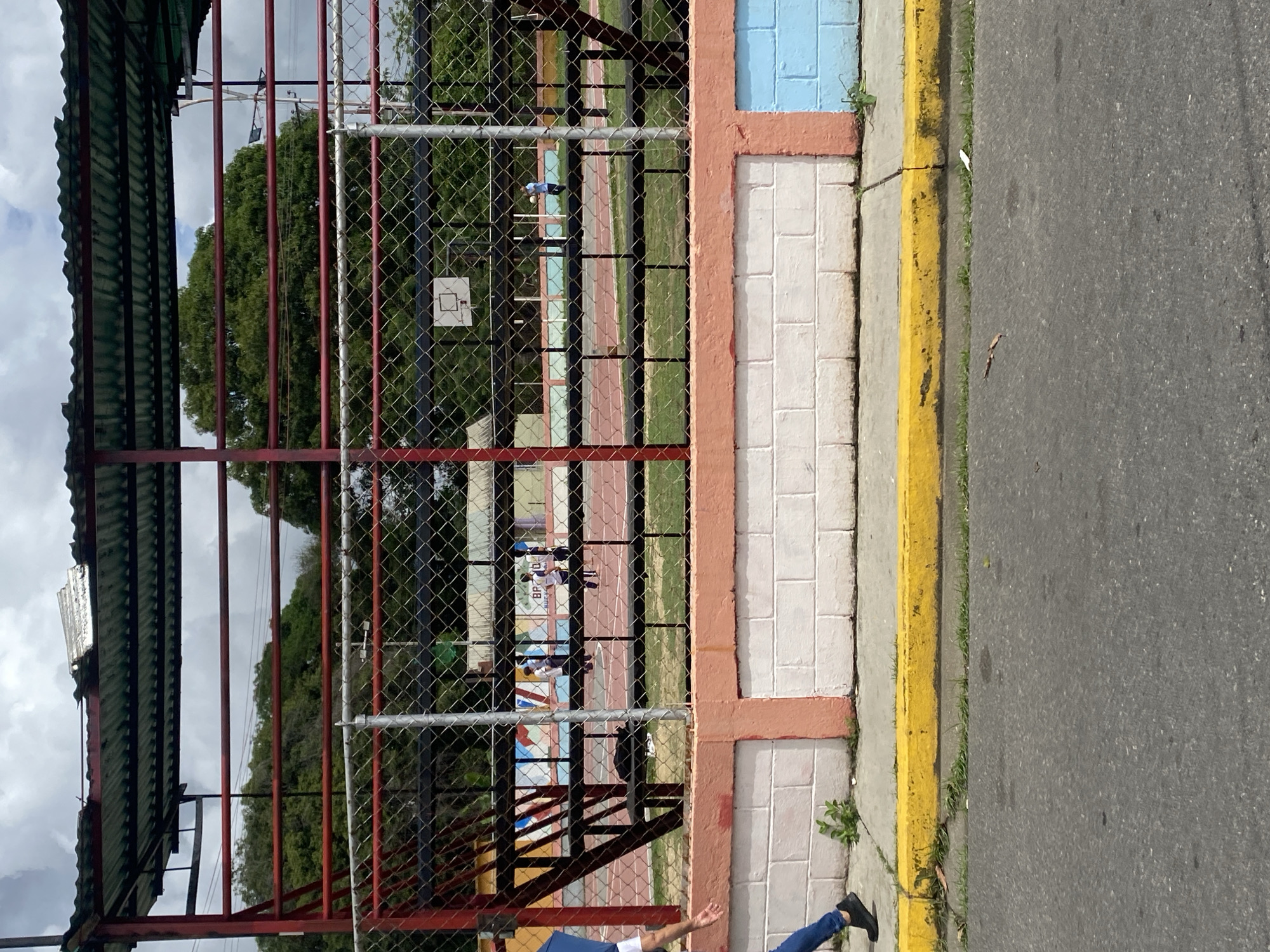 The image depicts an outdoor scene in Tinaquillo, Venezuela, next to a fenced sports facility with a track. The facility is enclosed by a chain-link fence. The fence is partially obscured by a red-painted metal structure, possibly a covered seating area. A basketball hoop is visible behind the fence. A section of painted brick wall with blue and pink trim is along the sidewalk. A sidewalk with a yellow curb borders the street. Part of a person wearing a blue shirt and jeans is visible in the bottom corner. The street is asphalt.