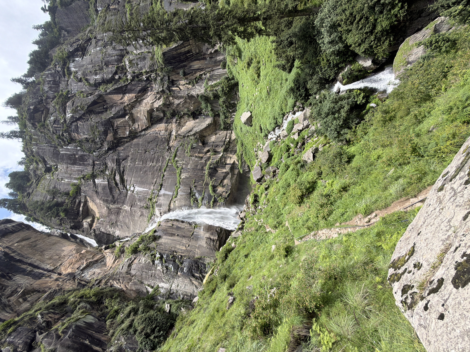 The image depicts the majestic Jogini Falls in Bahang, India, a multi-tiered waterfall cascading down a steep, rocky mountainside. Lush green vegetation blankets the slopes, interspersed with large boulders. A narrow, winding stone path with a metal railing is visible, leading upwards towards the falls. Several trees, including tall conifers, dot the landscape. The sky is partly cloudy. The scene is serene and natural, showcasing the raw beauty of the Himalayas. There are no people visible in the image.
