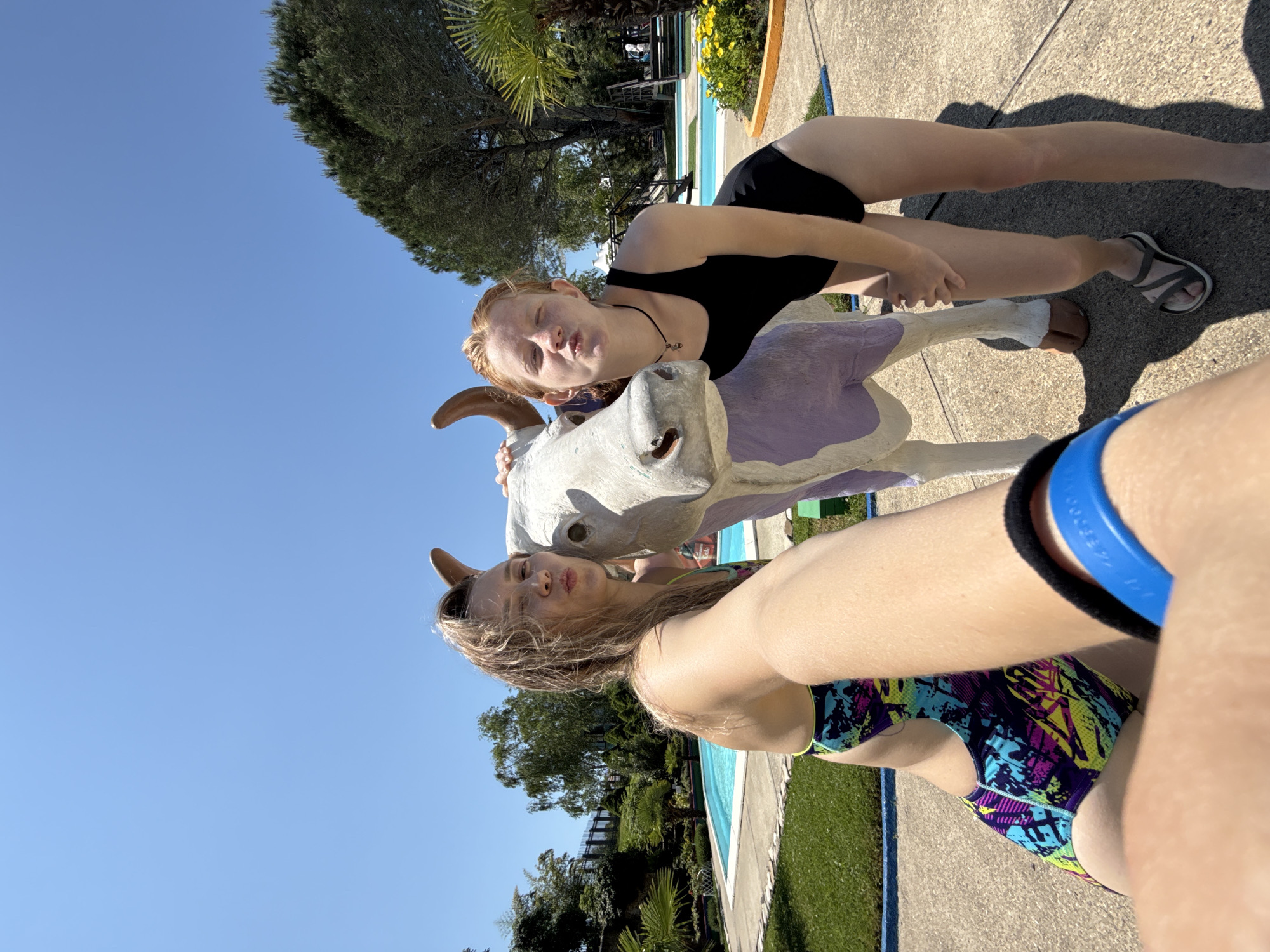 In Nebug, Russia, two young women pose for a selfie with a statue of a cow in an outdoor area. The woman on the left, with blonde hair, wears a colorful swimsuit and is positioned closer to the camera, leaning towards the cow. The woman on the right, with auburn hair and freckles, wears a black swimsuit and stands slightly behind the cow, looking at the camera. The cow statue has white and purple coloring and appears to be made of concrete. In the background, a swimming pool, lush green trees, and palm trees are visible under a clear blue sky, suggesting a warm, sunny day at a recreational facility or resort. The ground is paved with concrete and there is a patch of grass to the left of the pool.