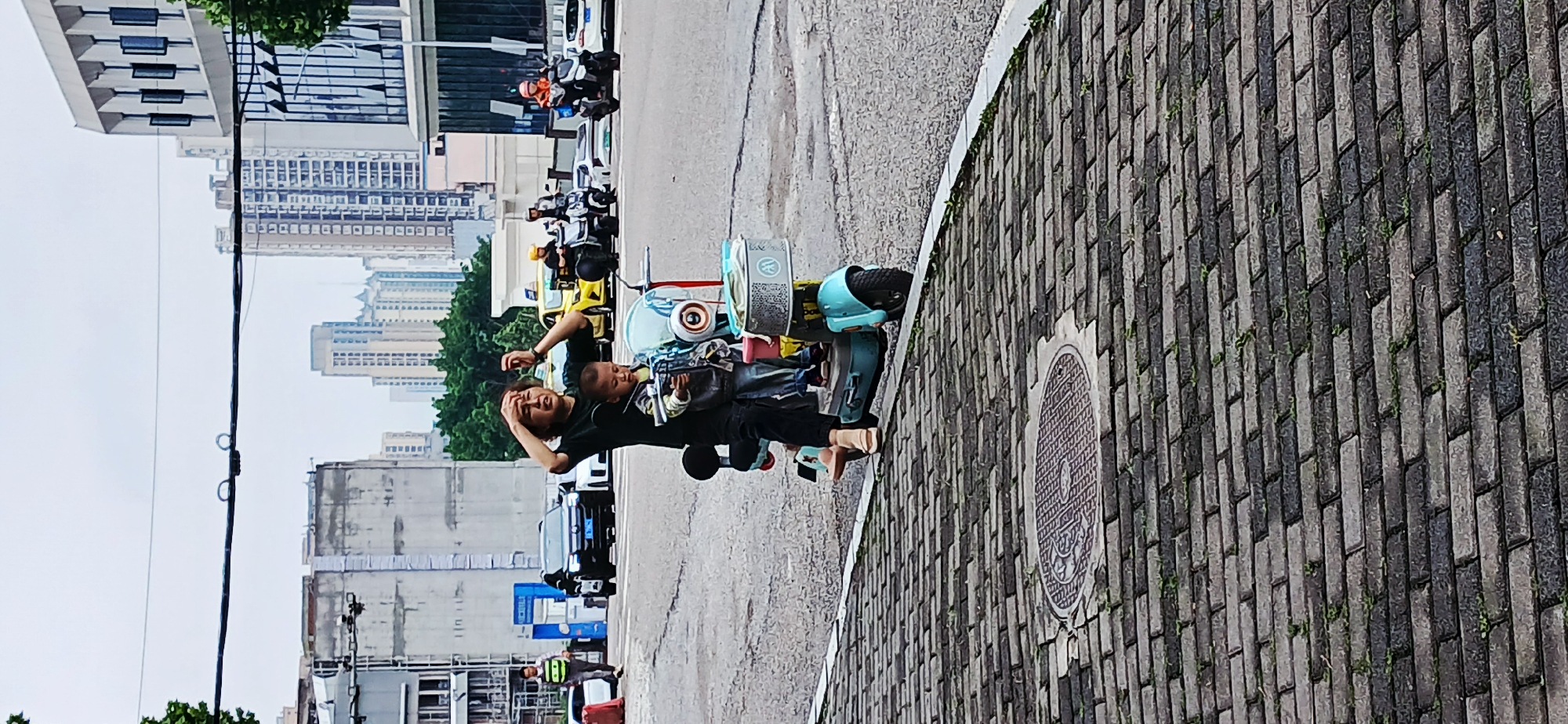 A man and a child are on a scooter on a street in Jiulongpo District, China. The man is wearing a black shirt and black pants. The child is sitting in front of him. There are other parked motorcycles and cars in the background. The cityscape is visible in the background, including buildings and trees. The street surface is paved with a section of patterned brick-like material and a section of asphalt. A manhole cover is visible on the paved brick section.