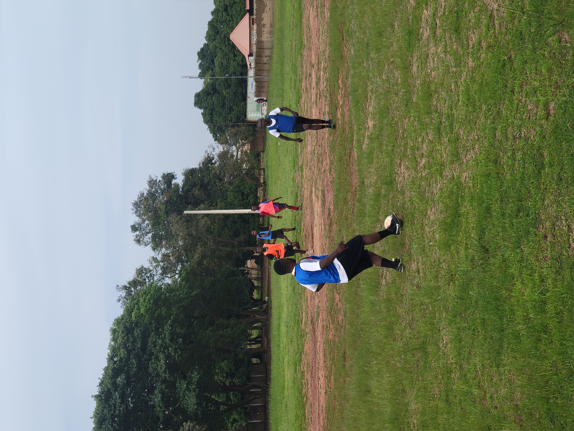 The image depicts a group of young men playing soccer on a grassy field in Jos, Nigeria. The scene is set outdoors with a clear sky, suggesting it is daytime and the weather is fair. The field is a mixture of green grass and bare earth, typical of a well-used sports ground. In the background, there are trees and some buildings, including a structure with a red roof and a shipping container, hinting at a semi-urban or developed rural setting.
The primary activity is a soccer game or training session. Several individuals are visible on the field, most of whom are wearing athletic attire. One person in the foreground is actively kicking a soccer ball. Another player in a blue and white jersey is in motion, seemingly participating in the game. Other individuals, some wearing bright vests like orange and pink, are scattered across the field, suggesting a practice drill or a casual match.
The people appear to be focused and engaged in the sport, with no clear indication of specific emotions other than concentration. The image captures a moment of athletic activity and camaraderie. The orientation of the image is rotated, with the sky appearing at the top and the ground at the bottom, which is a notable detail. No visible text is discernible in the image.