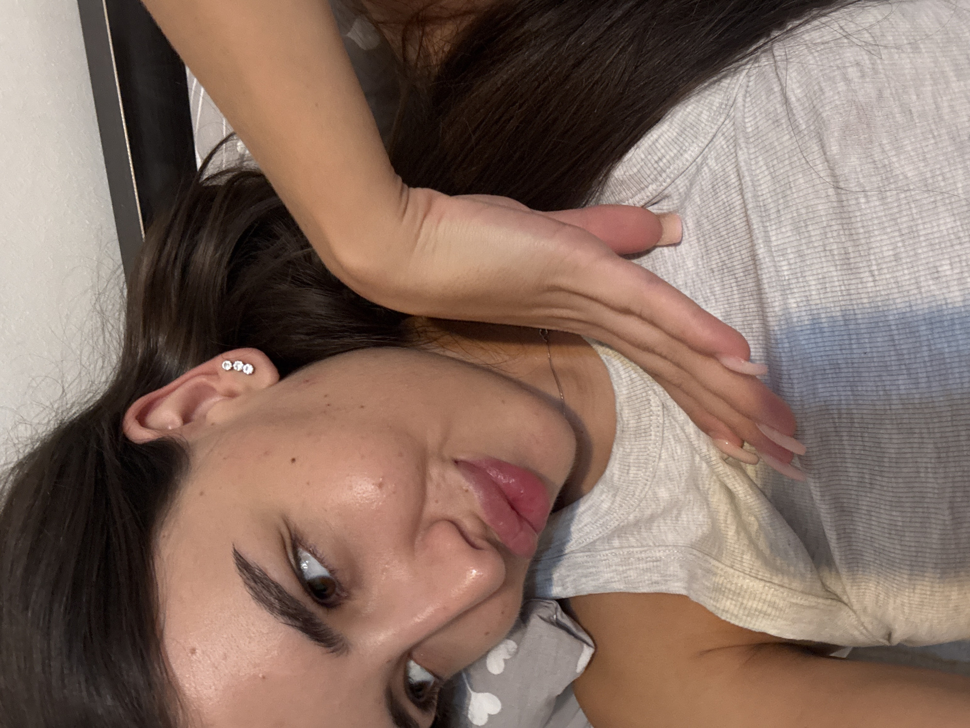 The image is a close-up, overhead shot of a young woman lying down, with her head tilted to her left. She has long, dark brown hair, and is wearing a light grey ribbed tank top. She has on three small, clear stud earrings in her left ear. Her right arm is bent, with her hand resting on her neck and shoulder, and her fingers extended. She has long, light pink nails. The background appears to be a bed with a grey and white patterned pillow and a dark headboard. The lighting is soft, and the overall impression is one of relaxation. There is no indication of the location within the city of Chișinău, Moldova.