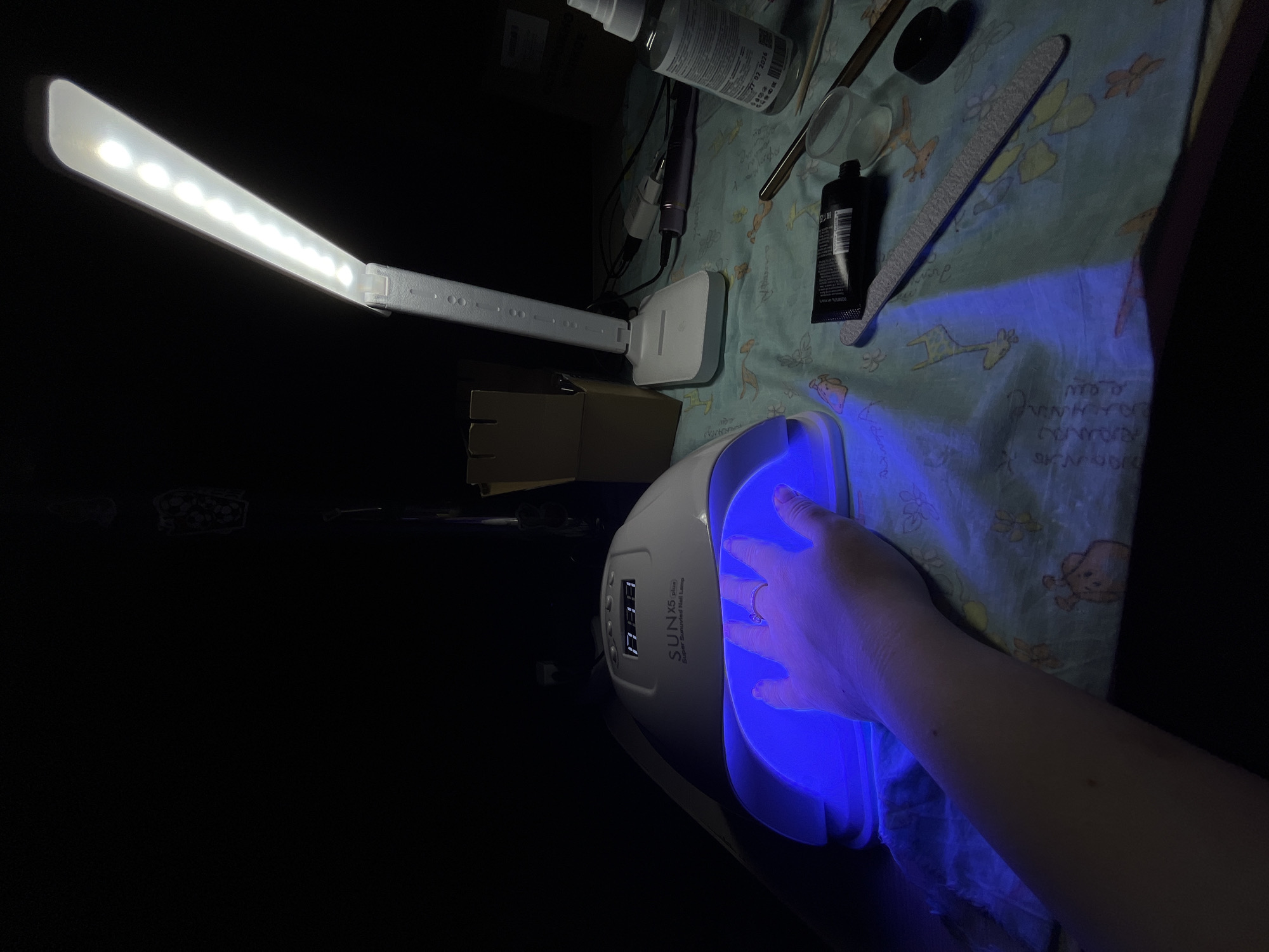 The image shows a manicuring setup on a patterned surface in dim lighting, possibly indoors. A person's hand is placed under a white SUN X5 plus Super Sunuvized Nail Lamp emitting a bright blue light. A white LED desk lamp illuminates the scene from above, with various manicure tools scattered around, including nail files, a tube of cream, and other small containers. The environment appears to be a personal or home setting. The location is specified as Novopokrovskaya, Russia, though there is no further evidence of such in the provided image.
