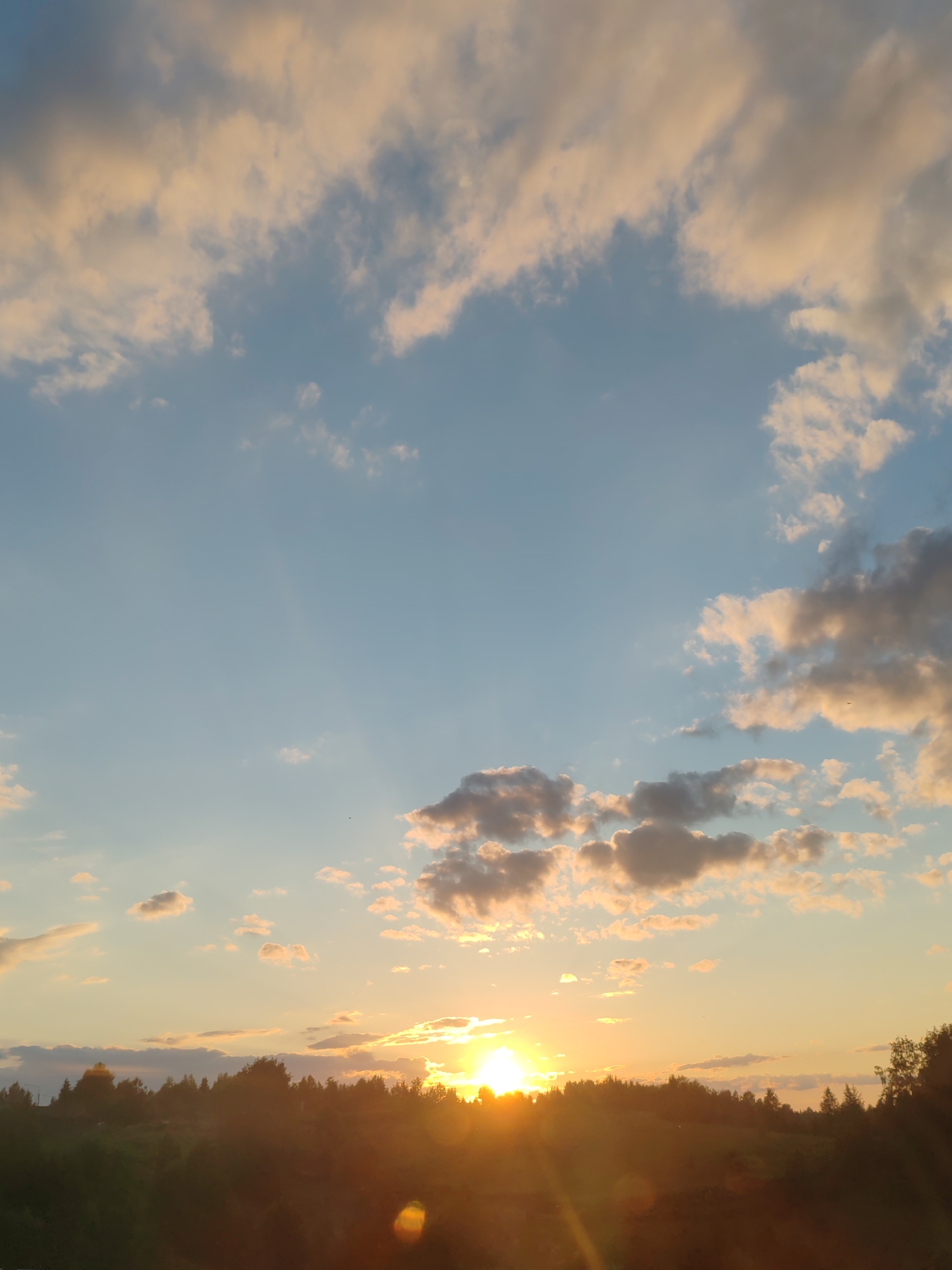 This is a photograph of a sunset over a treeline, taken in Чухлэм, Russia. The sky is a blend of blue and orange hues, with scattered clouds illuminated by the setting sun. The sun itself is a bright, glowing orb, partially obscured by the horizon. A dark band of trees runs across the lower portion of the image, silhouetted against the vibrant sky. There are no people or specific landmarks visible in the image.