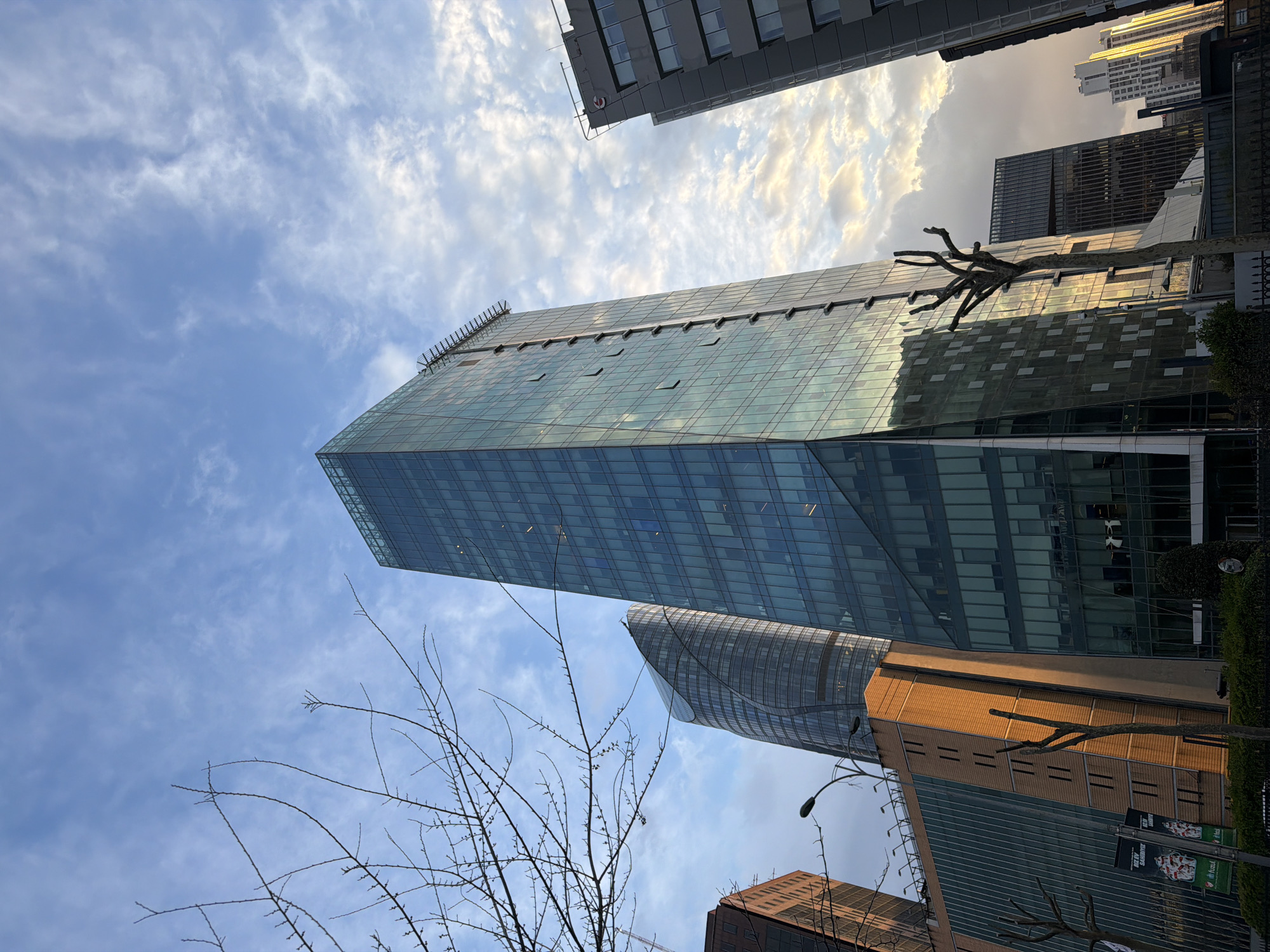 The image is an upward-looking perspective of modern architecture and an urban skyline in Sarıyer, Turkey. A tall, rectangular glass-facade building dominates the frame, reflecting the partly cloudy sky. Other buildings, including a cylindrical structure, are visible around the primary building. Bare tree branches are silhouetted in the foreground. The sky is a gradient of blue with scattered clouds illuminated by the sun.