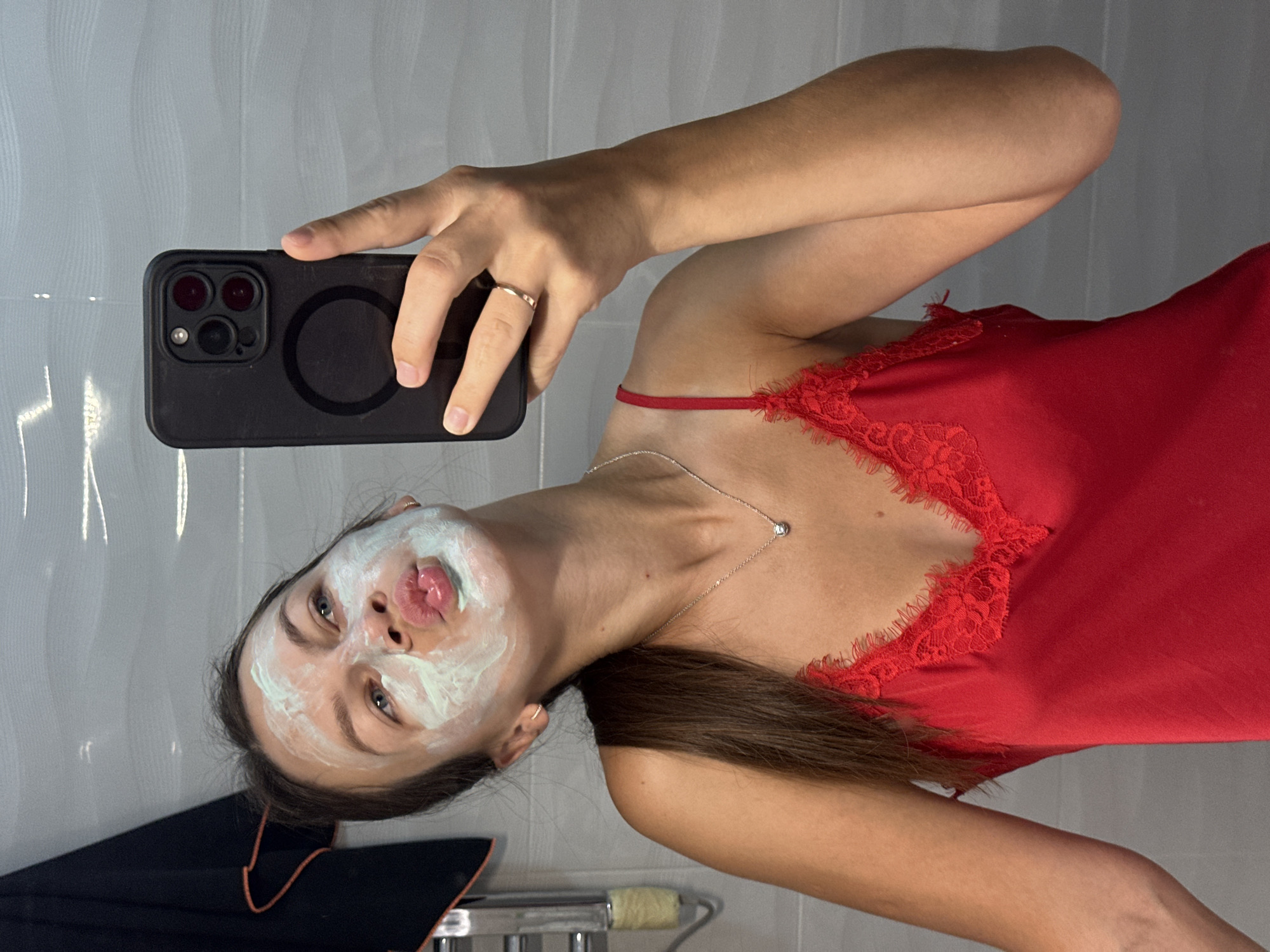 A woman with brown hair is taking a selfie in a bathroom. She has a light green face mask on her face and is puckering her lips. She is wearing a red camisole with lace trim and a silver necklace with a small diamond pendant. She is holding a black smartphone in her right hand. The background is a tiled wall. The image does not provide any information about the city of Boreti, Montenegro, or any specific location.