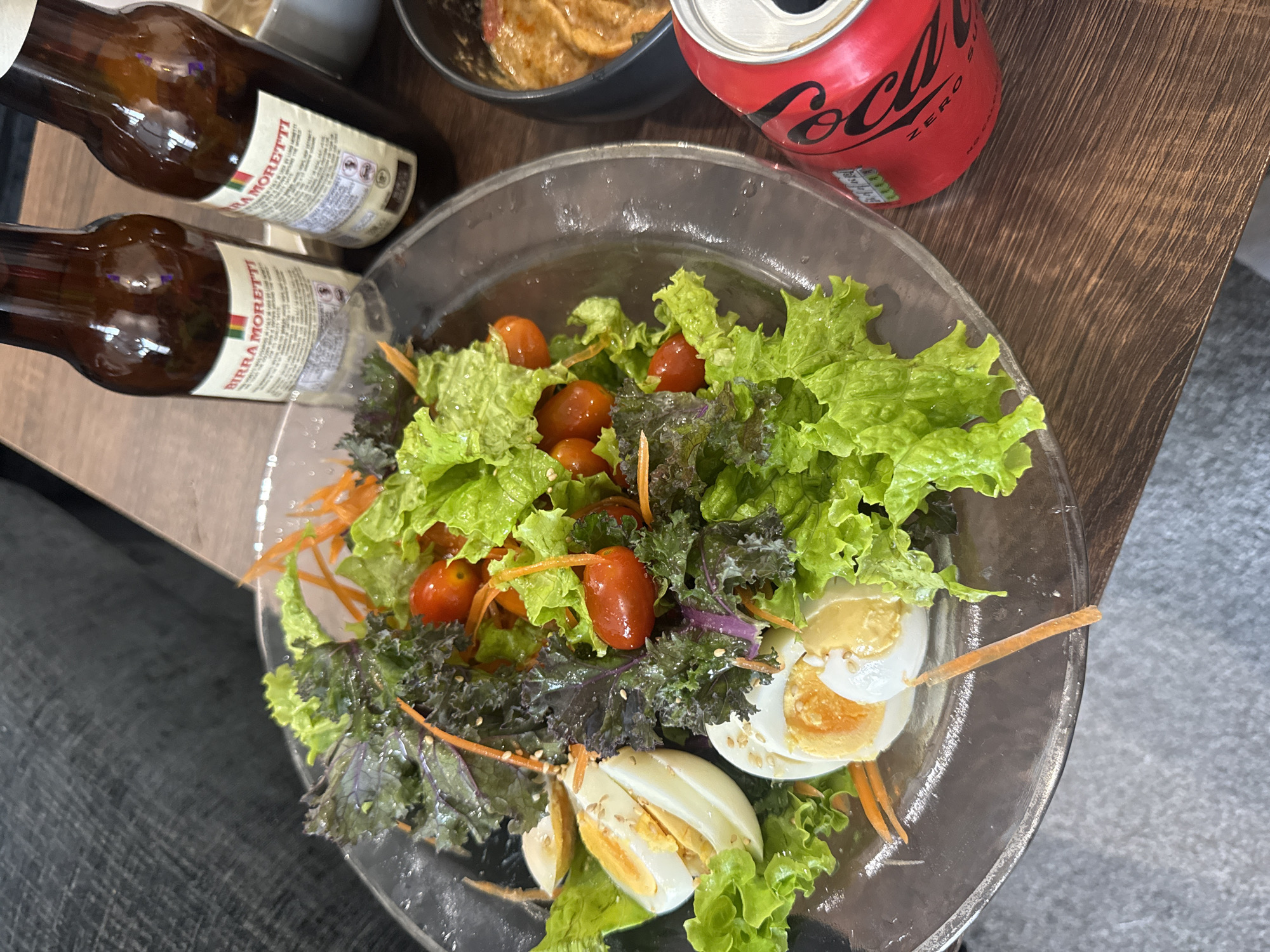 A close-up, overhead shot shows a salad and beverages on a wooden surface. The salad, served in a clear glass bowl, features lettuce, cherry tomatoes, shredded carrots, kale, and hard-boiled eggs. Two bottles of Birra Moretti beer and a red Coca-Cola Zero can are also visible. The background is blurred, but suggests an indoor setting with a textured grey surface and a dark fabric. The scene appears to be set on a table, and the lighting suggests it might be daytime, though no specific location in Throop, United Kingdom, can be determined from the image.