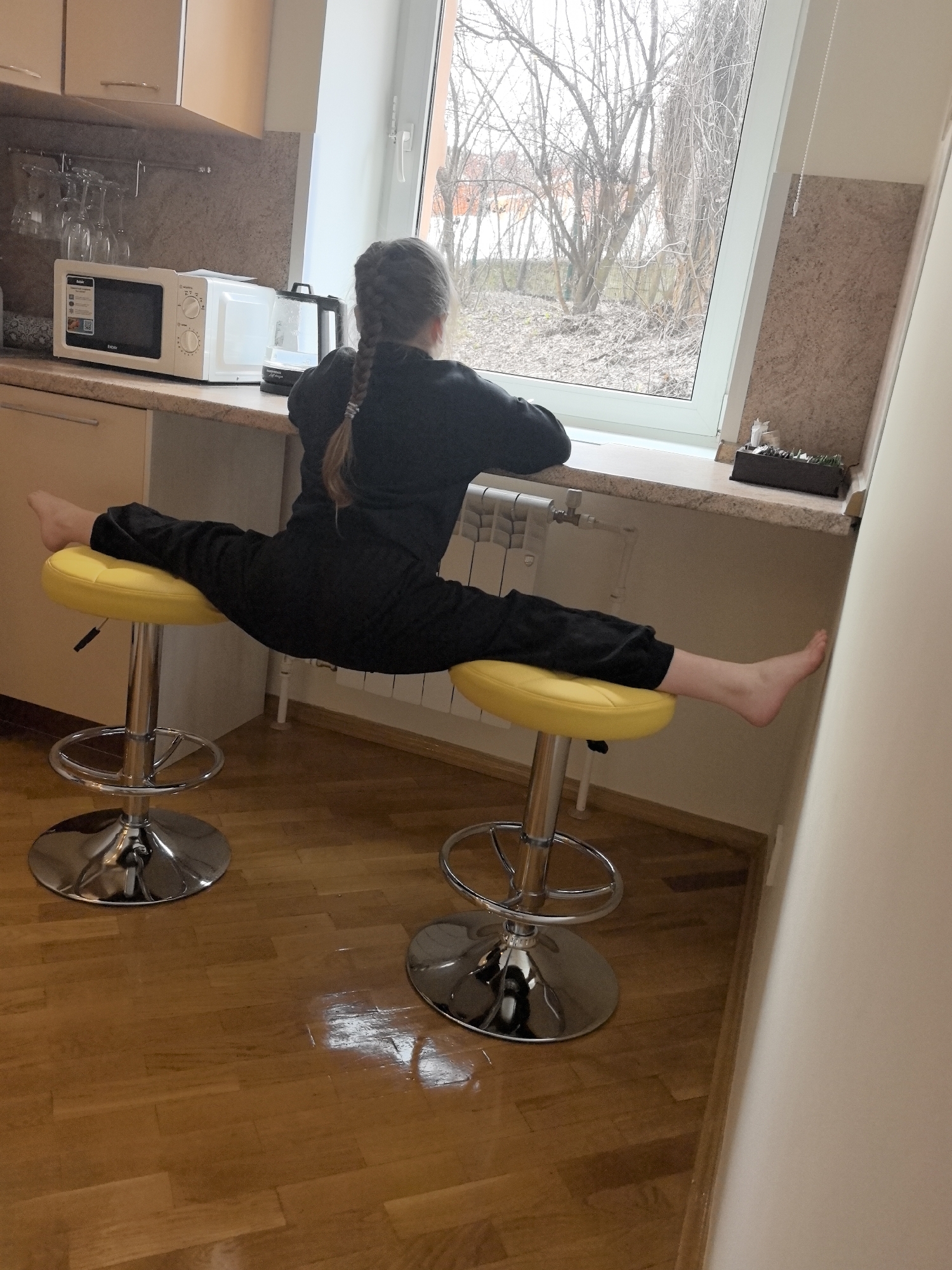 A girl in a black jumpsuit is performing a split on two yellow bar stools in a kitchen. A window behind her shows trees outside, possibly in Nizhny Novgorod, Russia. The kitchen includes cabinets, a microwave, and a radiator.