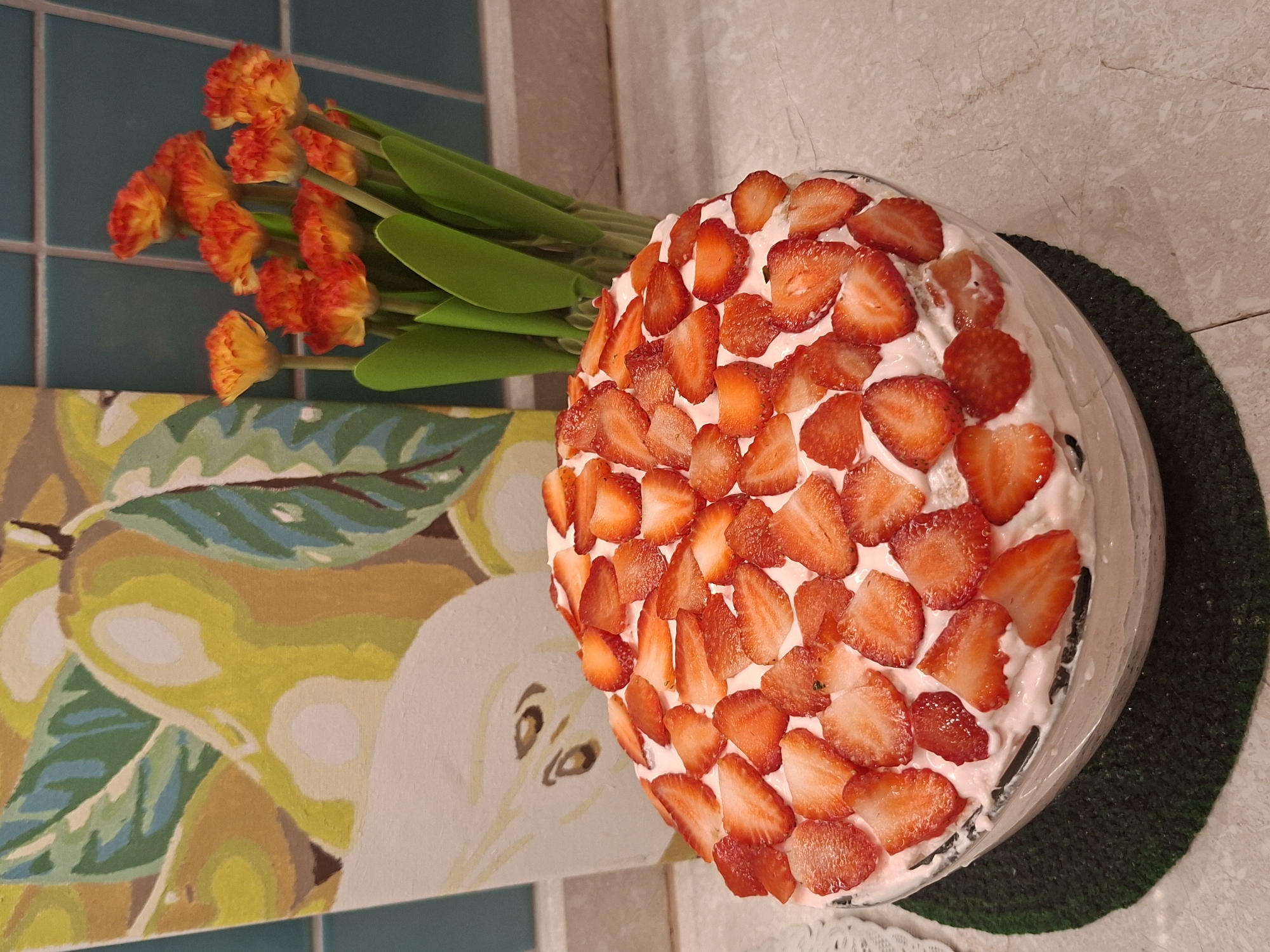An overhead shot features a circular cake topped generously with sliced strawberries and a pink frosting, resting on a clear glass stand. The cake is placed on a dark green mat. In the background, a painting depicting pears and leaves hangs on a tiled wall. To the right of the cake, a vase holds a bouquet of orange and yellow flowers with green stems. The scene is set on a light-colored countertop with a subtle marble pattern. The lighting suggests an indoor environment. The image appears to be a still life of a dessert, possibly in a home kitchen or dining area, with decorative elements adding to the composition.