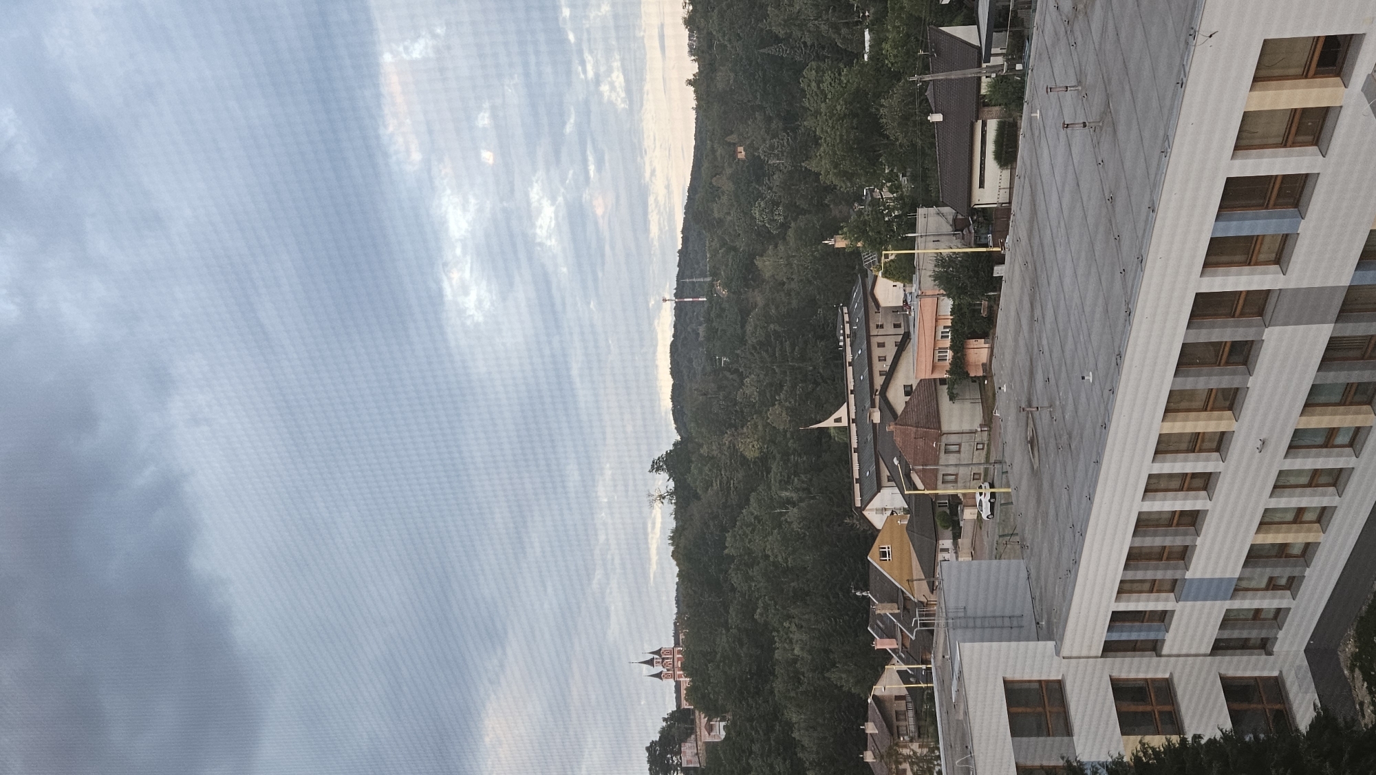 The image shows a view of a town and surrounding hills on a cloudy day. In the foreground, a modern building with rows of windows is visible. Behind it, there are several houses with pitched roofs. The middle ground features a densely wooded hill. On top of the hill, a distinctive pink building with multiple spires can be seen, along with a red and white tower structure further to the right. The sky is overcast with layers of gray clouds, some of which are lighter and may be illuminated by the sun. A fine mesh pattern is visible across the entire image, suggesting it was taken through a screen or net. The scene depicts a residential area nestled against a natural landscape. This view is consistent with the location of Kalvária in Prešov, Slovakia, where a similar church complex is situated on a hill overlooking the town.