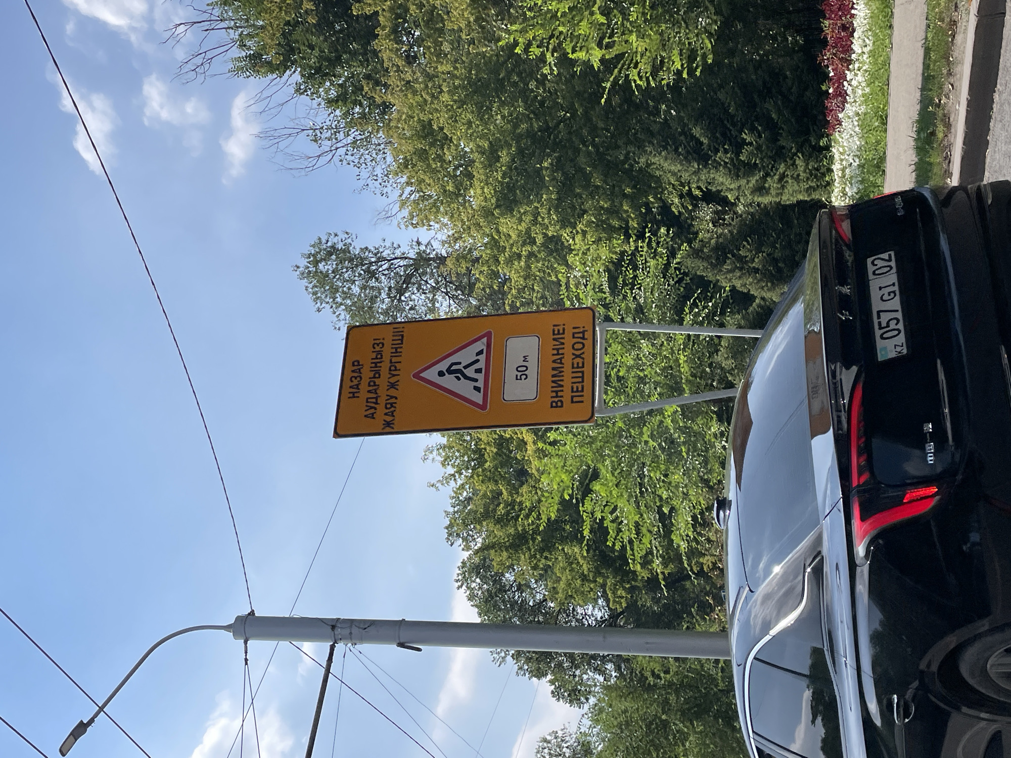 A black sedan is parked on a street in Almaty, Kazakhstan. A traffic sign indicating a pedestrian crossing is visible in the background. The sign has text in Kazakh and Russian, and a symbol of a pedestrian. The car has a license plate with the text "KZ 057 GI 02". The scene is set against a backdrop of trees and a blue sky with clouds.