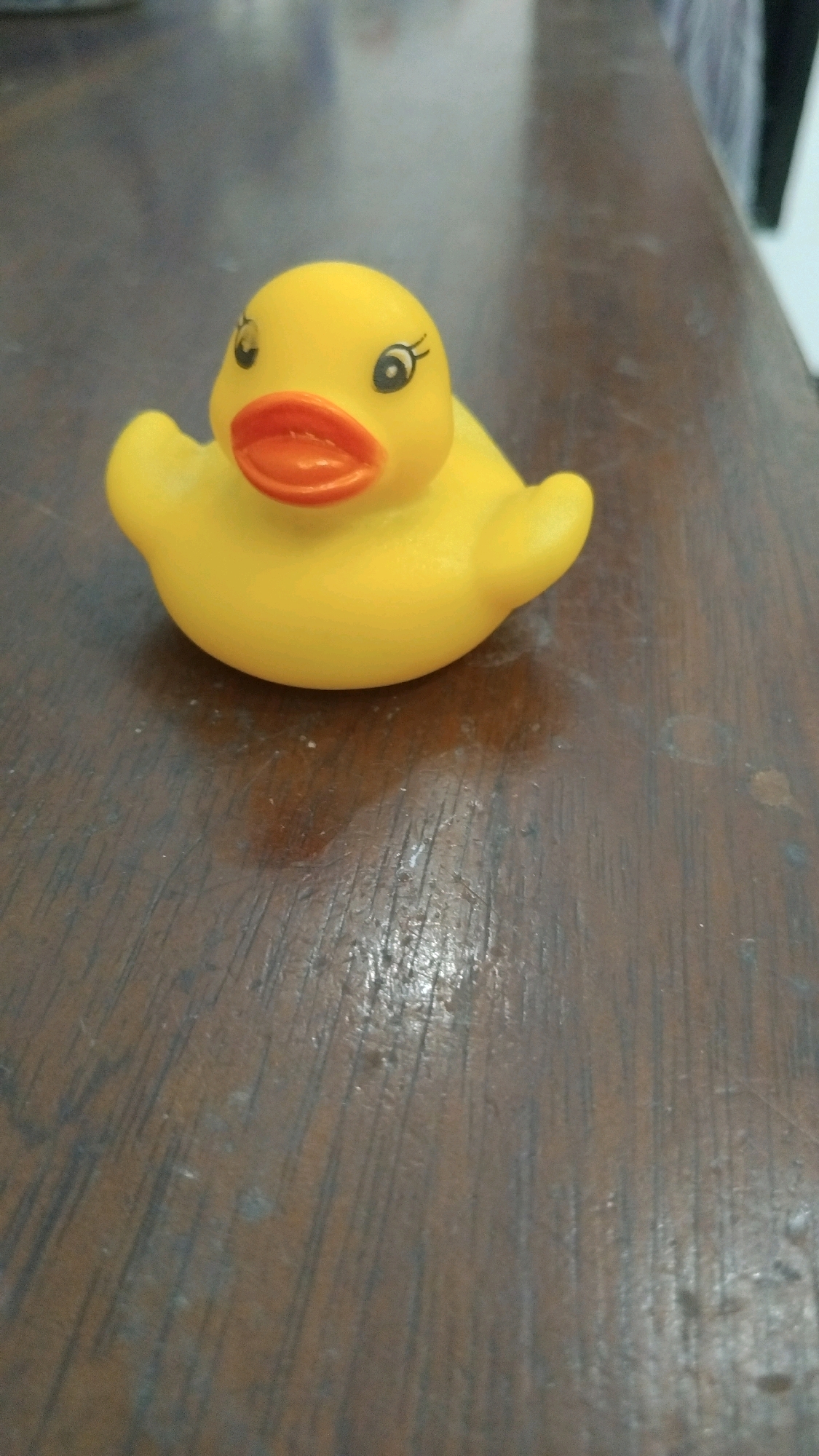 A close-up, eye-level, slightly angled shot shows a yellow rubber duck toy positioned on a brown wooden table. The duck is facing left, with its orange beak slightly open, and painted-on eyelashes. The table surface has a visible wood grain pattern and appears worn, with some smudges and lighter-colored marks. The background is out of focus, with muted colors and indistinct shapes, suggesting an indoor setting. There is no information about the location in the city of Kebon IX, Indonesia, present in the image.