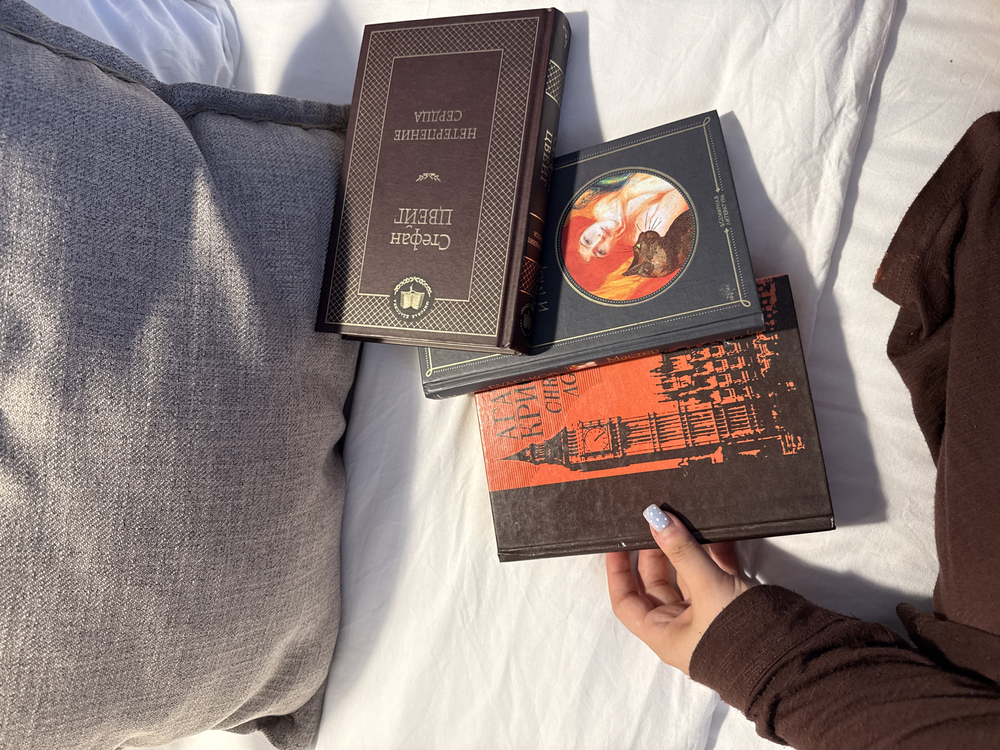 Three books are arranged on a white, rumpled surface, likely a bed or sofa, with a textured grey pillow in the background. A person's hand, with light blue nail polish featuring white polka dots, is holding the left book. The book on the left has a dark brown cover with an orange illustration of a clock tower, likely Big Ben, and some Russian text. The middle book is dark blue with a circular illustration of a woman with red hair and a black cat. Russian text is visible on its cover. The top book is dark brown with gold lettering and a decorative border, also featuring Russian text. The overall scene is a close-up view of these books and a hand, illuminated by natural light creating shadows and highlights. There is no identifiable connection to Starokorsunskaya, Russia, in the visual information provided.