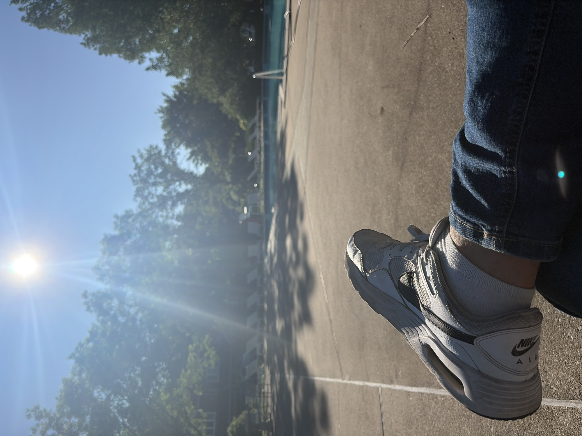 The image depicts a person sitting outdoors in Edison, New Jersey, United States. The person is wearing blue jeans, white socks, and white Nike Air shoes. The location appears to be near a pool, which is partially visible in the background. The sun is shining brightly, creating lens flare. Trees surround the pool area. The ground appears to be concrete or paved.