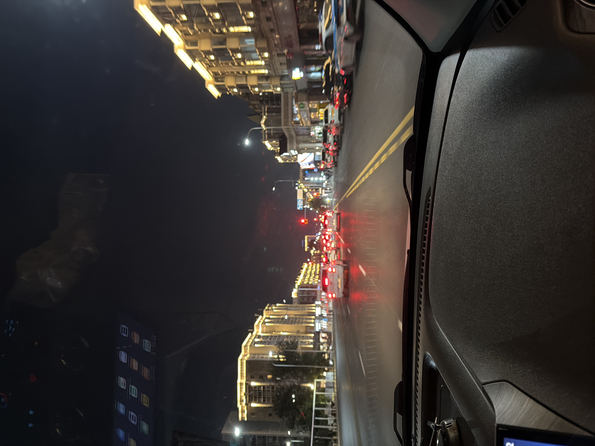 The image is a point-of-view shot from inside a vehicle driving on a city street at night. The street is wide with multiple lanes and yellow lane markings. The buildings lining the street are modern and illuminated with warm-toned lights. Several cars are on the road, their headlights and taillights creating streaks of light. The sky is dark, indicating it is nighttime. A portion of the vehicle's dashboard is visible in the foreground. The reflection on the windshield shows abstract patterns and some blurred shapes. This scene is consistent with a street view in Astana, Kazakhstan, particularly along one of the main thoroughfares, potentially during evening hours when the city's architecture is highlighted by artificial lighting.