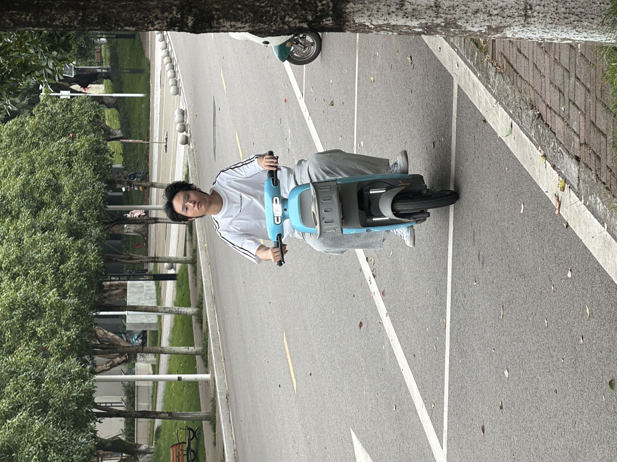 A young man with dark hair and glasses, wearing a white t-shirt with black stripes on the sleeves and gray sweatpants, is riding a light blue electric scooter down a paved road. He is looking straight ahead with a neutral expression. The scooter has a gray basket attached to the front. In the background, there are trees lining the sidewalk, and a row of white spherical bollards. To the right of the man, another green and white scooter is parked. The road is marked with white lines and a yellow directional arrow. The scene appears to be in an urban park or residential area.