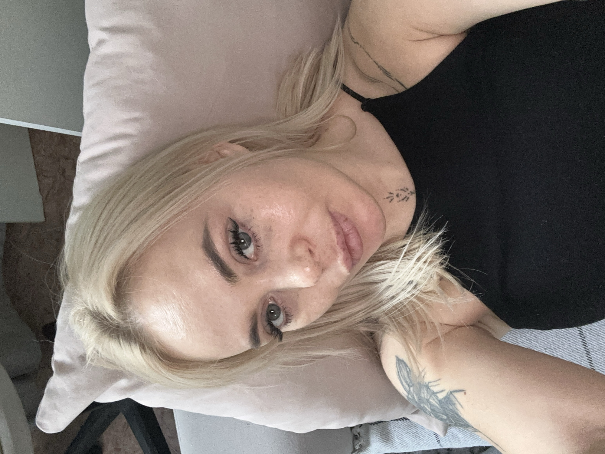 A woman with blonde hair is lying on a pink pillow. She is wearing a black tank top and has a tattoo on her neck and arm. The background includes furniture and a wall. The location is Высокий, Russia.