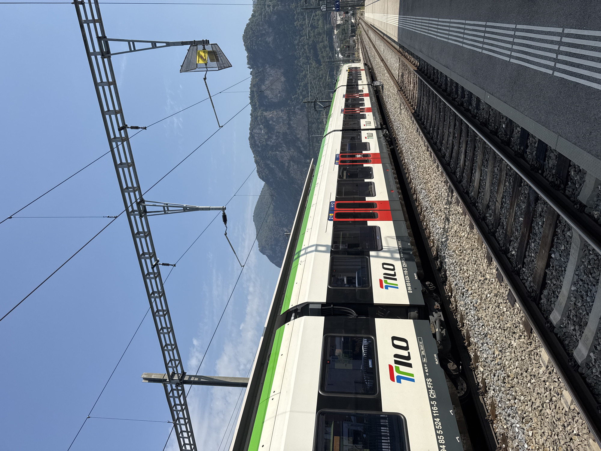 A white and green train with red doors is stopped at a station platform. The train has "TILO" branding on its side. The train tracks are visible, with gravel ballast. A concrete platform with tactile paving runs alongside the tracks. In the background, there are mountains covered in trees. The sky is clear and blue. The location is the train station in Mendrisio, Switzerland.