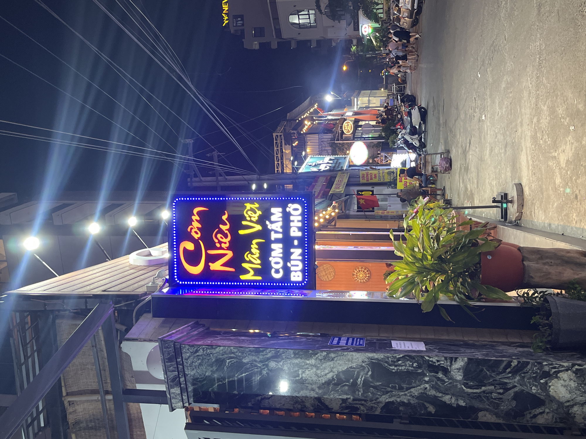 Here is a detailed description of the image:
**Setting:** The image is taken at night in Bac Nha Trang Ward, Vietnam. It appears to be a commercial area with restaurants and shops lining the street.
**People:** Several people are present in the image, mostly towards the back. They are likely patrons of the various food establishments. Some individuals can be seen walking along the sidewalk or are seated at tables in front of restaurants.
**Objects:**
* **Signage:** A prominent, brightly lit sign for "Com Niêu Mâm Việt" is in the foreground. Smaller signs and storefronts are visible along the street.
* **Vehicles:** Motorbikes are parked along the side of the road.
* **Street Furnishings:** Potted plants are near the foreground, potentially marking the entrance to a business.
**Environment:**
* The street is illuminated by streetlights and the glow of businesses.
* The overall atmosphere is lively and energetic.
* Power lines stretch across the sky above the street.