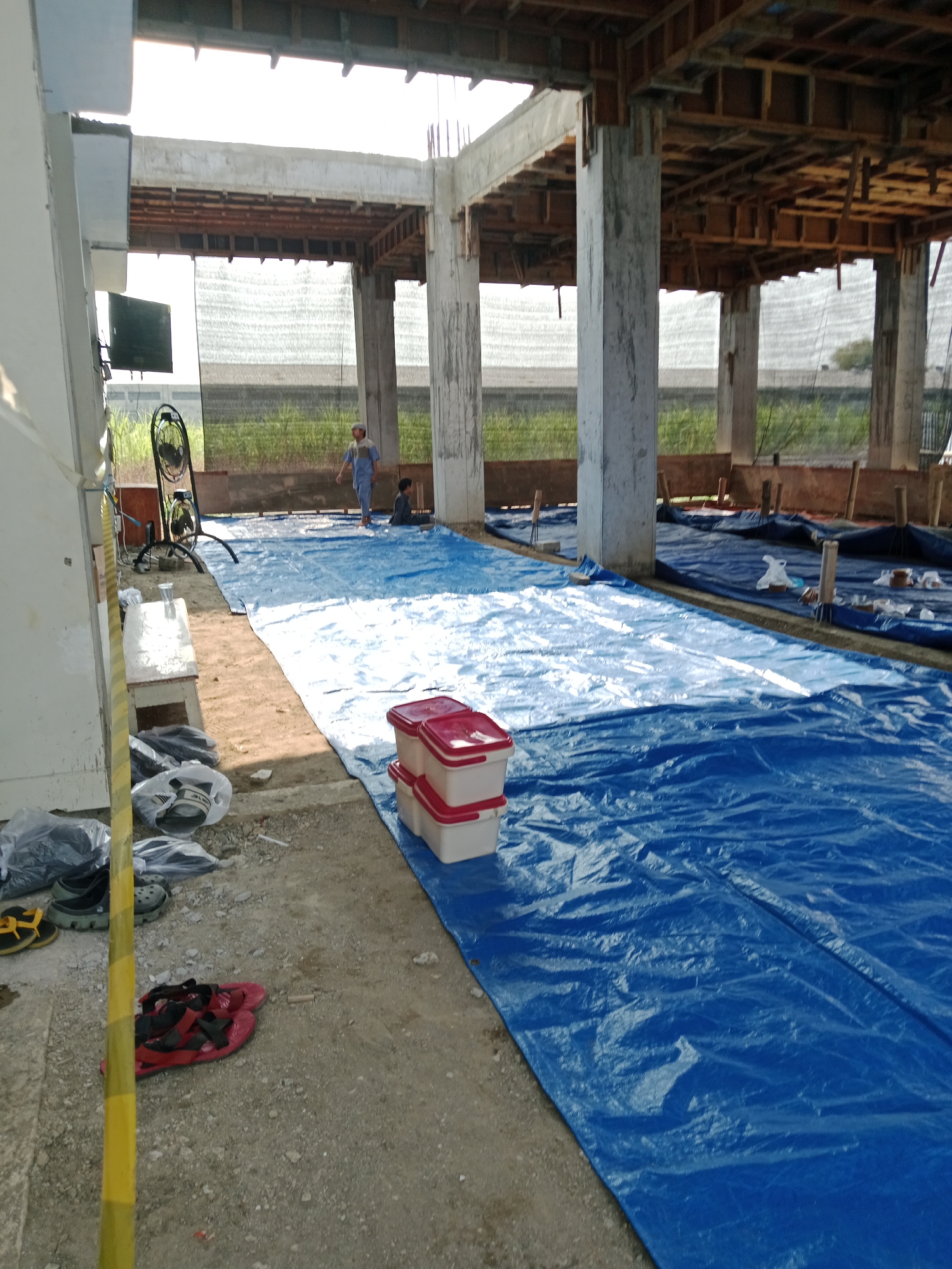 The image depicts a construction site in Kediri City, Indonesia. A large blue tarp covers a significant portion of the ground, extending from the foreground into the background. Several concrete pillars support an unfinished structure overhead, with wooden framing visible. In the middle ground, two individuals are present on the blue tarp. One, wearing a light-colored cap and blue clothing, is walking. Another person is seated on the tarp. To the left of the image, a white bench is situated on the sandy ground, next to a black and silver oscillating fan. Various items, including shoes and plastic bags, are scattered on the ground near the bench. In the foreground, red sandals are placed on the dirt. A stack of red and white plastic containers is positioned on the blue tarp. The overall scene suggests ongoing construction work under a bright, possibly sunny, sky.