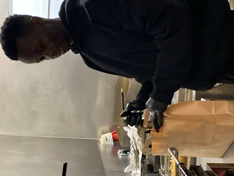 A person in a black jacket and gloves packs a brown paper bag. The person is leaning over a stainless steel counter in what appears to be a kitchen. Foil-wrapped food and cooking equipment, like fry baskets, are visible next to the bag. A white electrical outlet is also on the counter. The lighting is bright, and the room seems clean and functional.